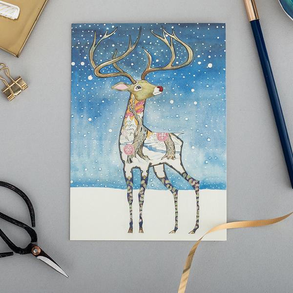 Illustration of a whimsical reindeer with decorative patterns on its body, displayed on high-quality Rudolph Cards from The DM Collection, against a snowy background on a craft table with scissors and ribbon.