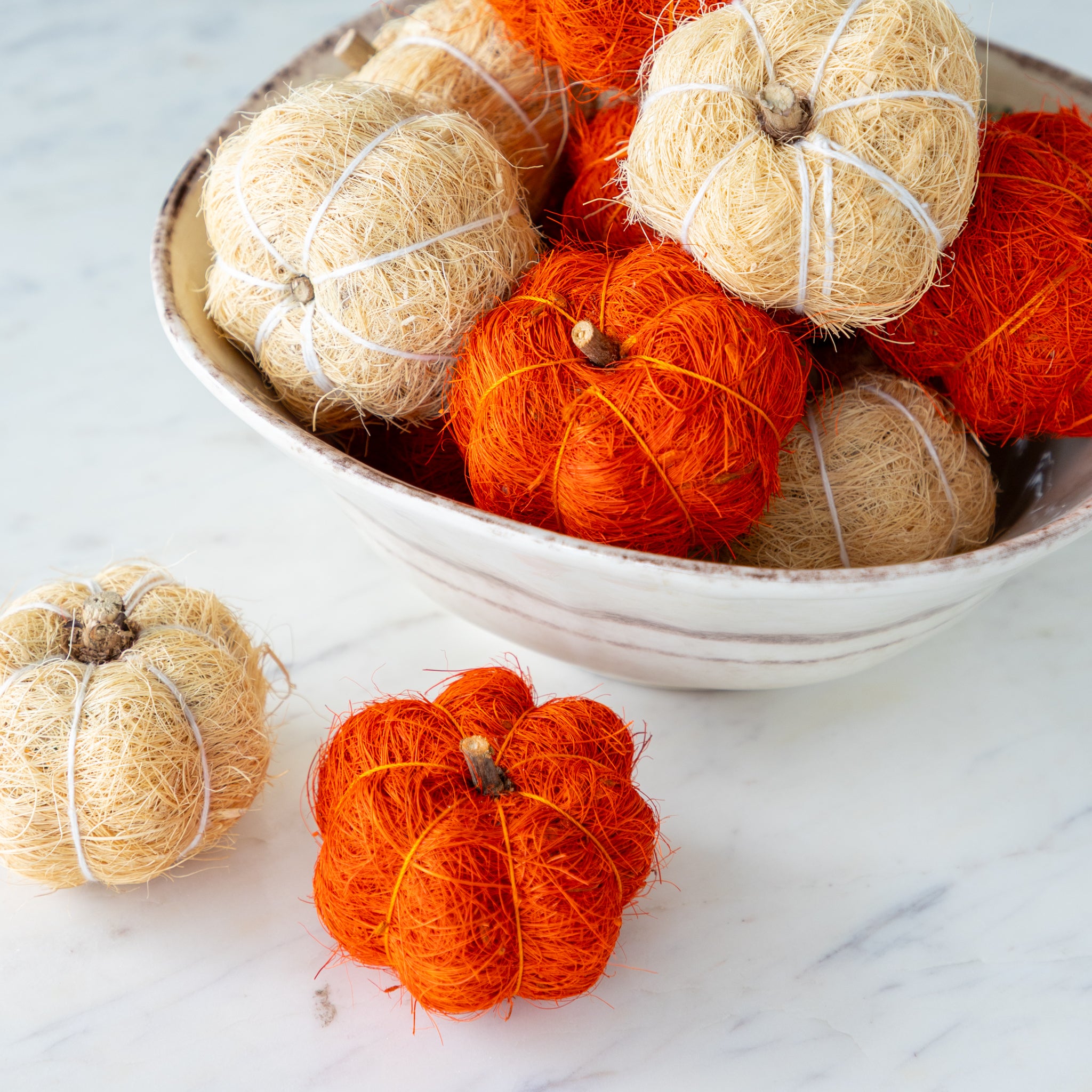 Coco fiber decorative pumpkins in burnt orange and cream in a bowl on a marble table.