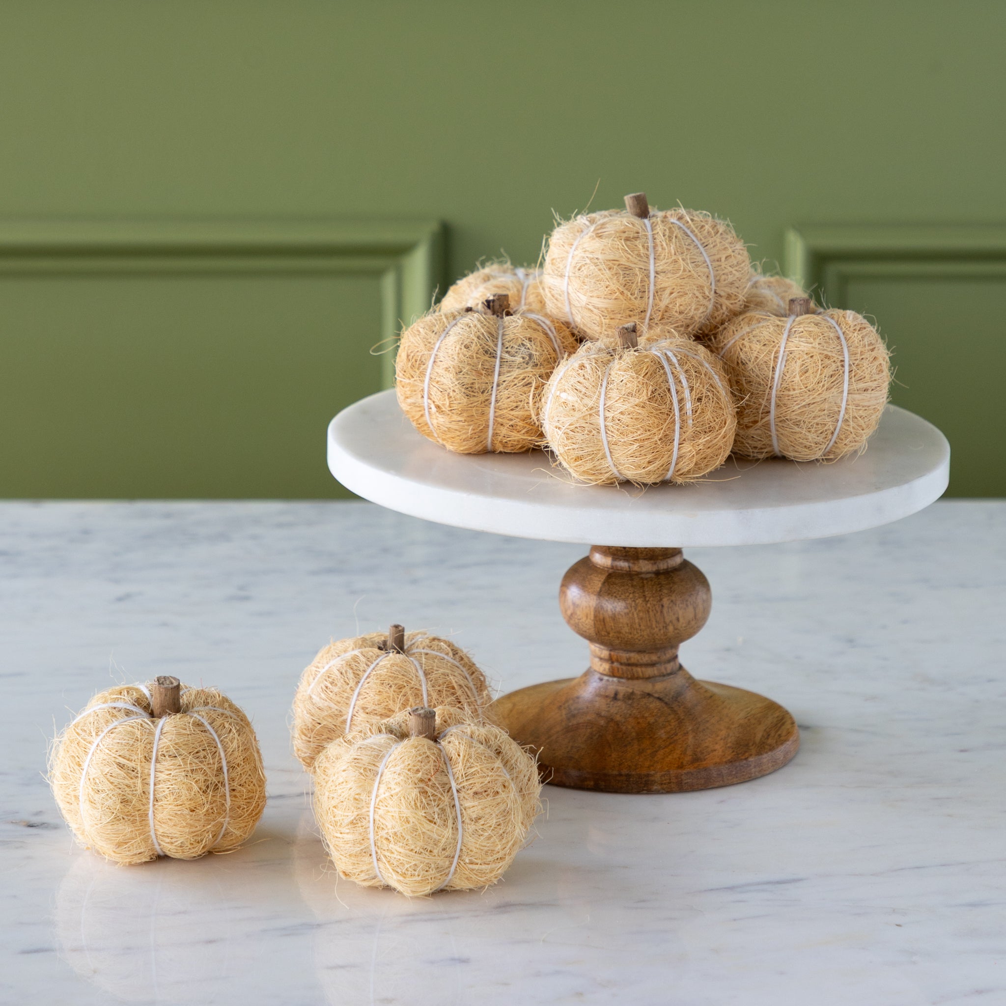 Cream coco fiber pumpkins on a white cake stand with a wooden base against a green wall.
