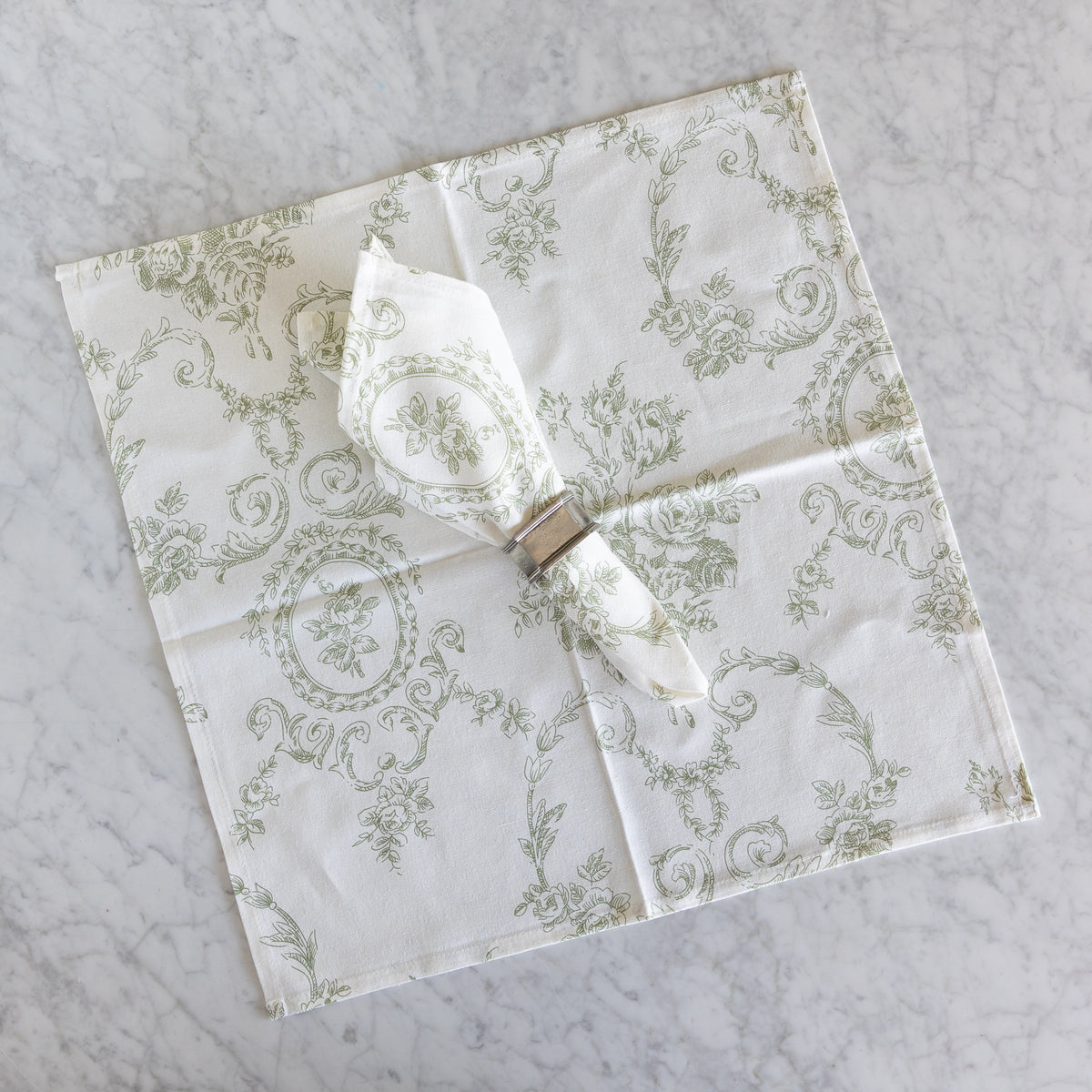 A square white napkin with a sage green toile pattern, folded and displayed on a light gray marble surface with a matching napkin ring holding a folded white napkin.