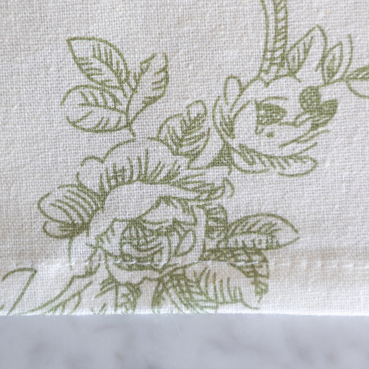A close-up of a white woven fabric with a sage green floral line pattern, resting on a light gray marble surface.