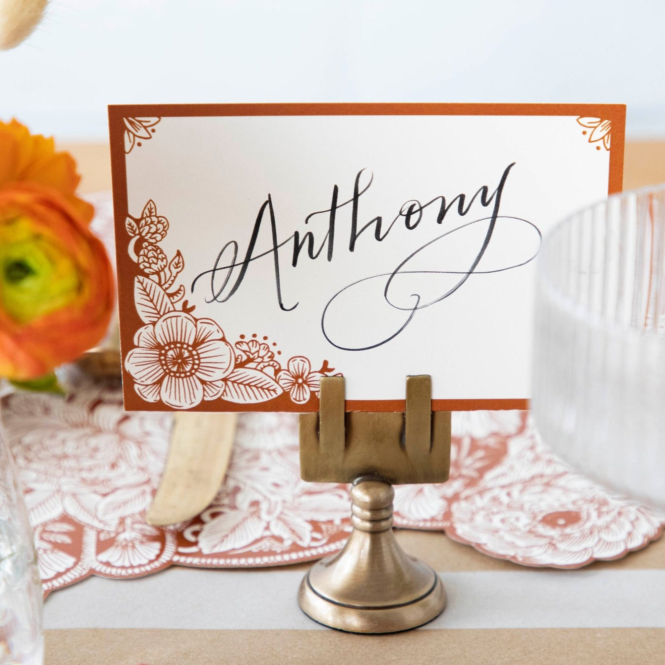 Close-up of a Brass Place Card Holder on an elegant fall-themed table setting, holding a card labeled &quot;Anthony&quot;.