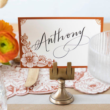 Close-up of a Brass Place Card Holder on an elegant fall-themed table setting, holding a card labeled &quot;Anthony&quot;.