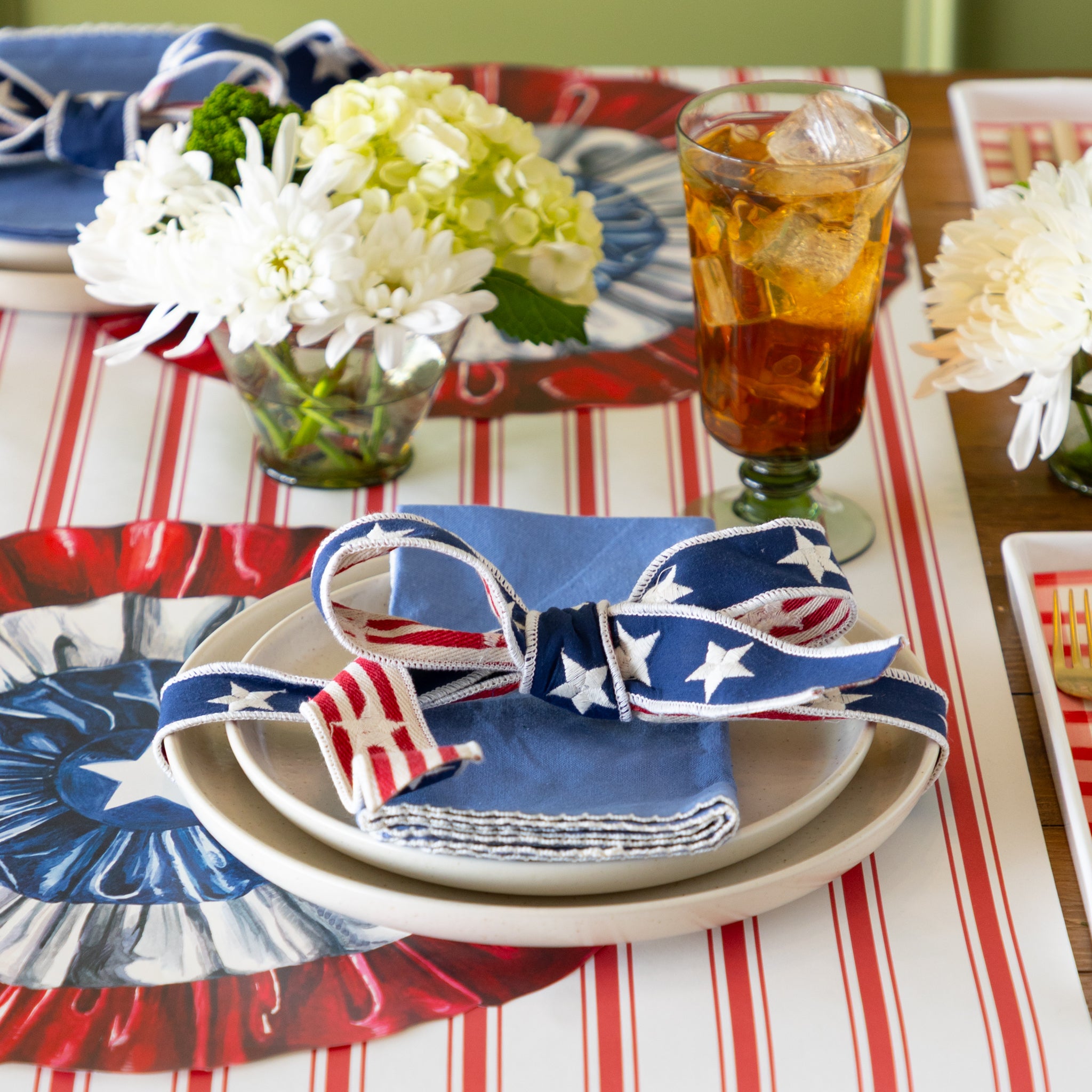 Table setting with American flag-themed ribbon tied around a napkin and plate on a table with flowers and a drink on a red and white striped table runner.