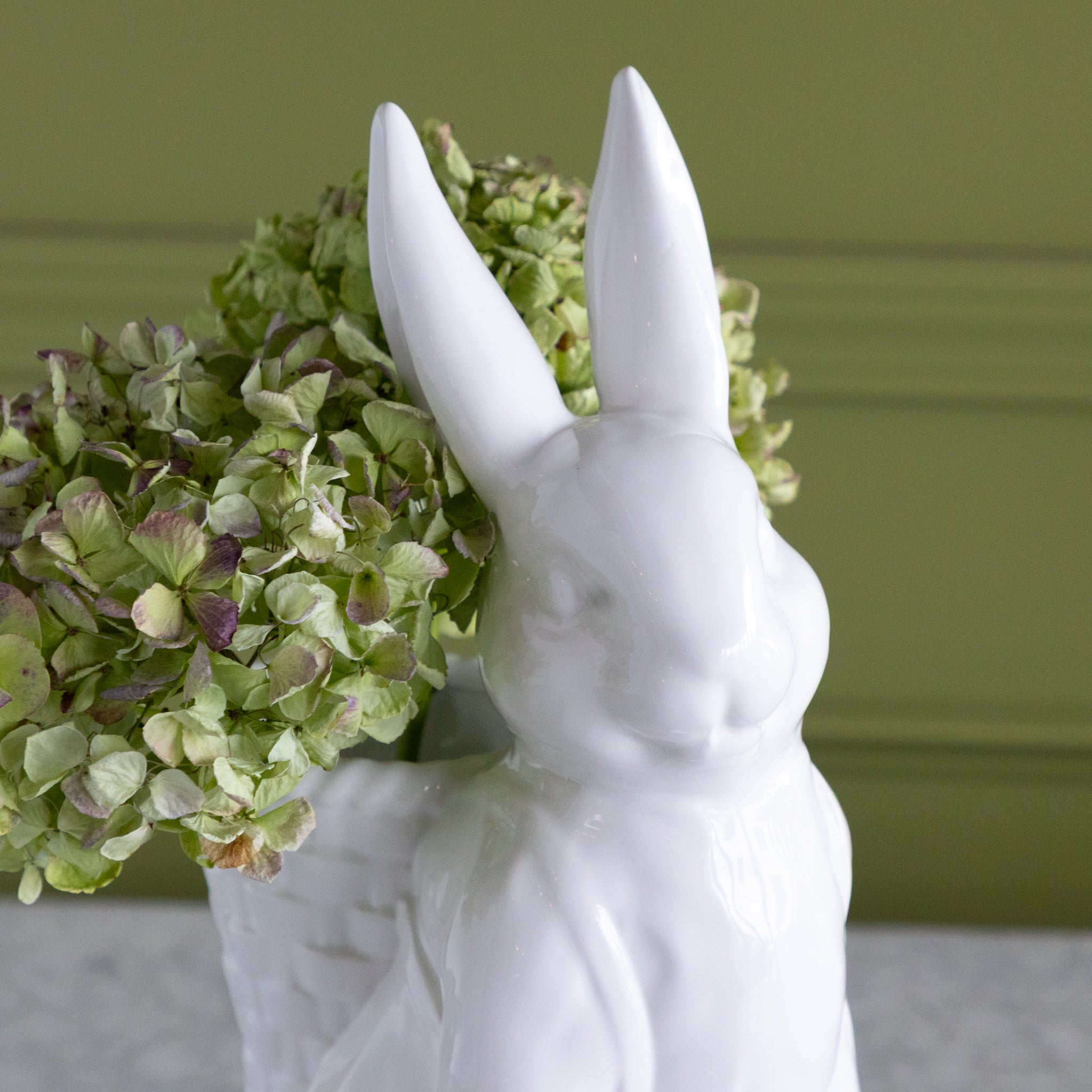 White rabbit-shaped vase with green hydrangeas in its basket against a green wall.