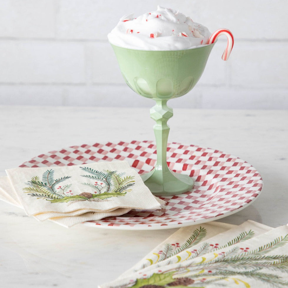 A table with a stack of Christmas Sprigs Cocktail Napkins and a stack of Christmas Sprigs Guest Napkins, fanned out next to a red and white plate and a cup of hot chocolate.