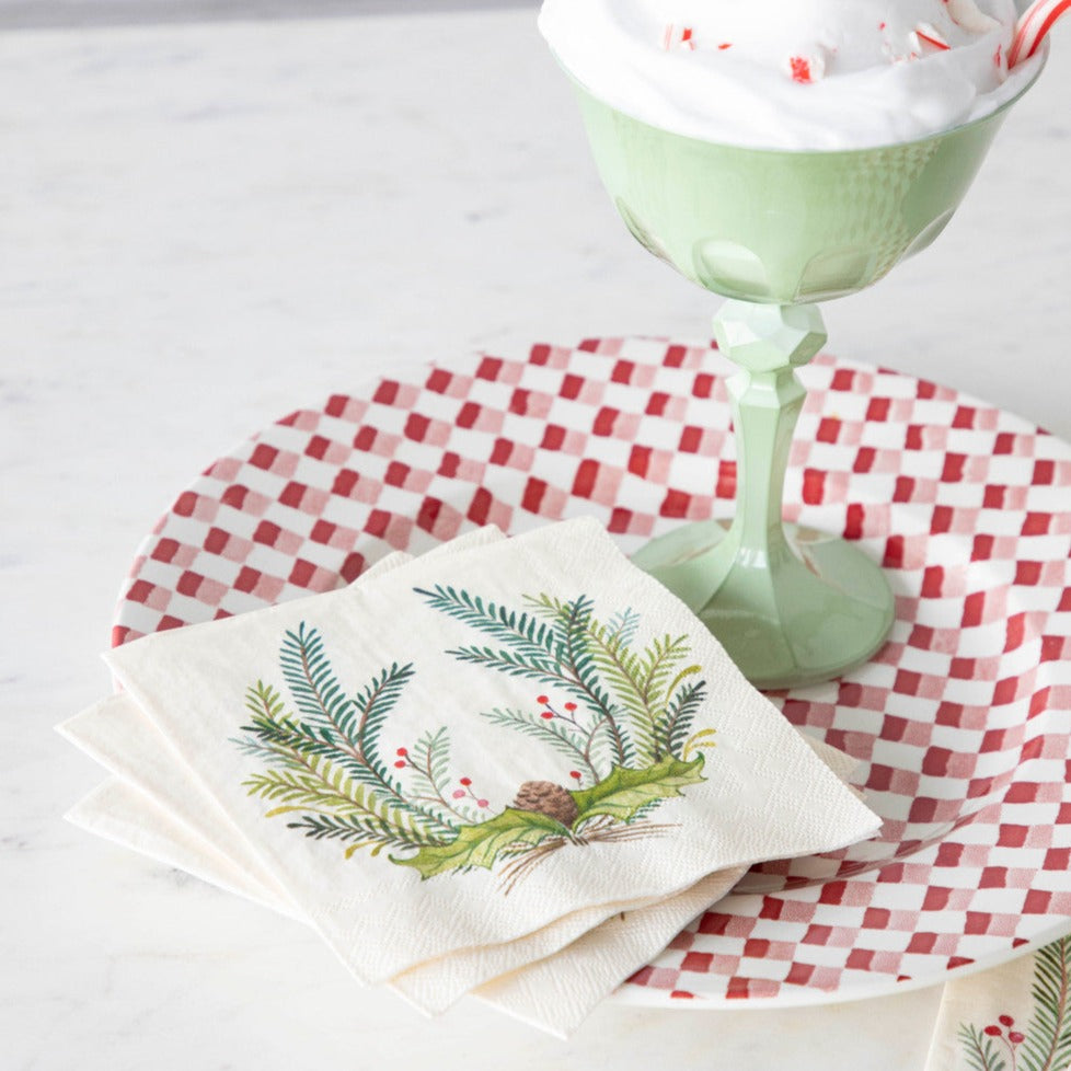 Three Christmas Sprigs Cocktail Napkins fanned out on a red and white plate with a cup of hot chocolate.