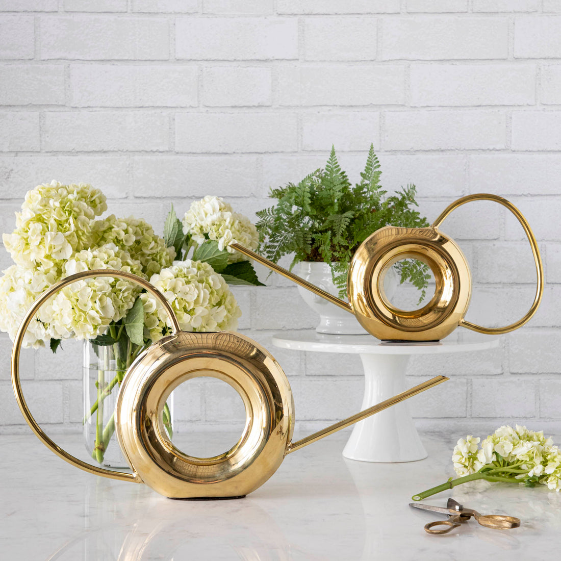 Two Accent Decor Modern Watering Cans with brass finish on a table next to flowers.