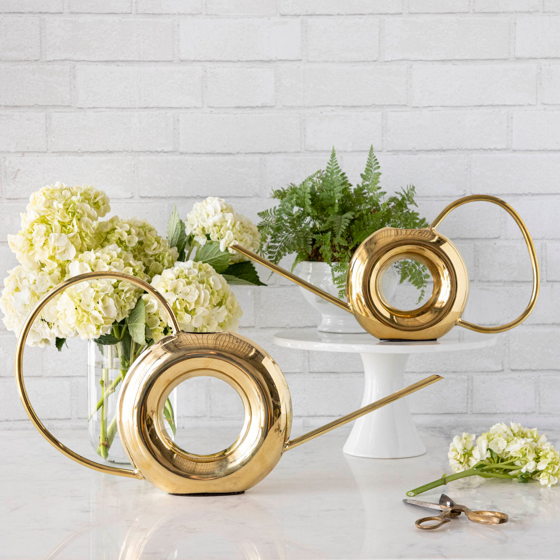 Two Accent Decor Modern Watering Cans with brass finish on a table next to flowers.