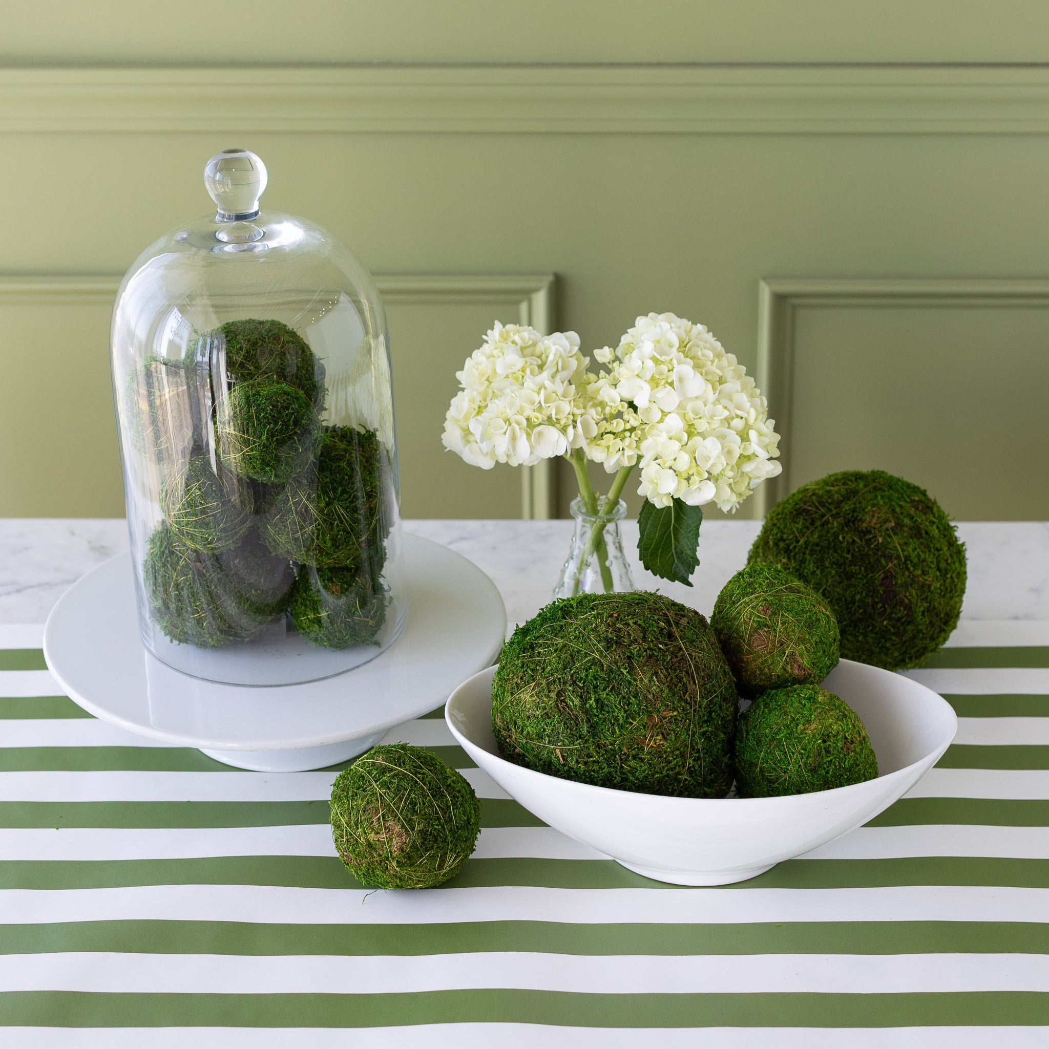 Moss balls displayed in a white bowl on a table with a striped tablecloth, with a glass cloche containing more moss balls in the background, alongside a vase with flowers.