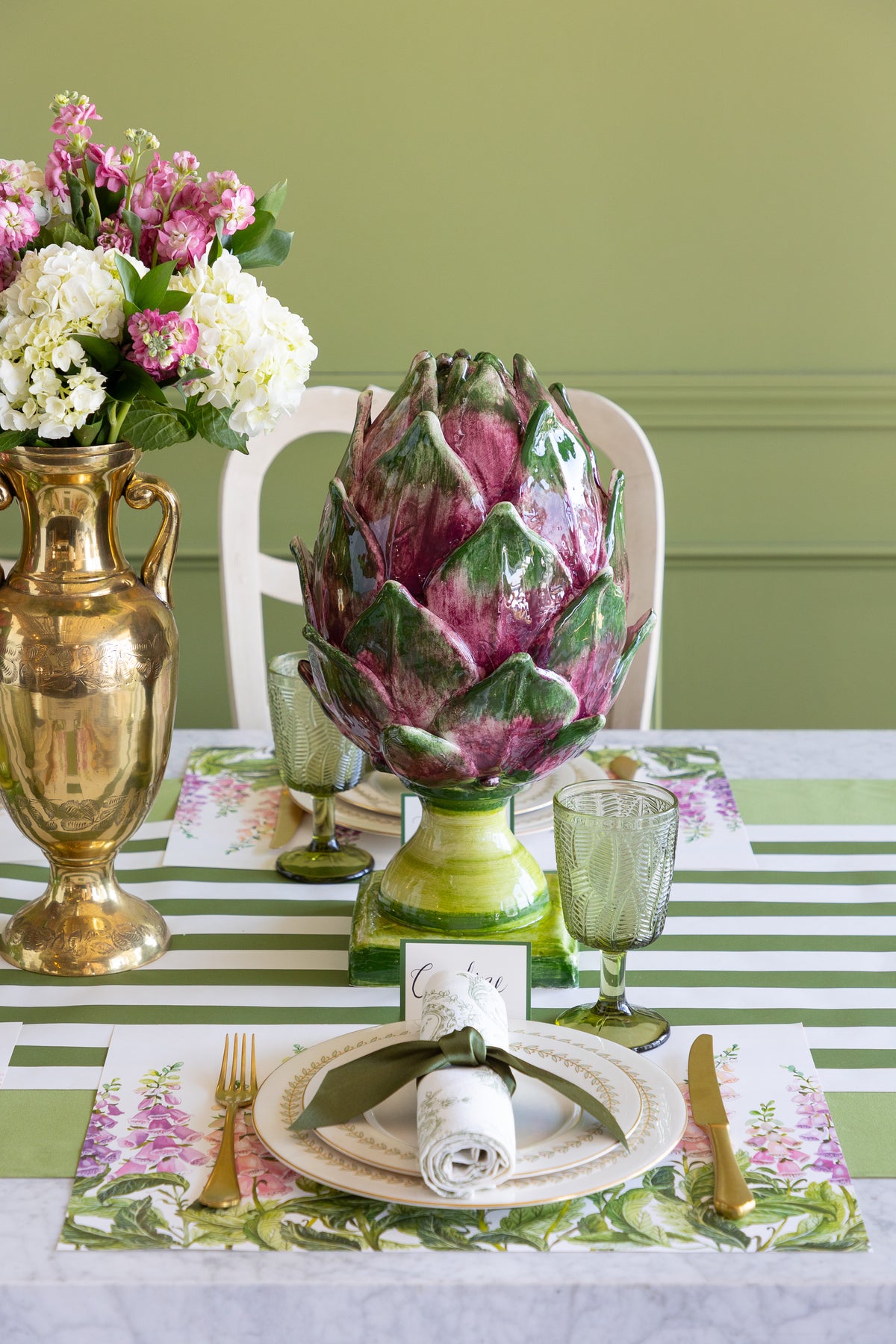 A styled dining table setting with a ceramic artichoke centerpiece in a green vase, a gold urn with flowers, green glass goblets, floral placemats, and a green-and-white striped tablecloth against a sage green wall.