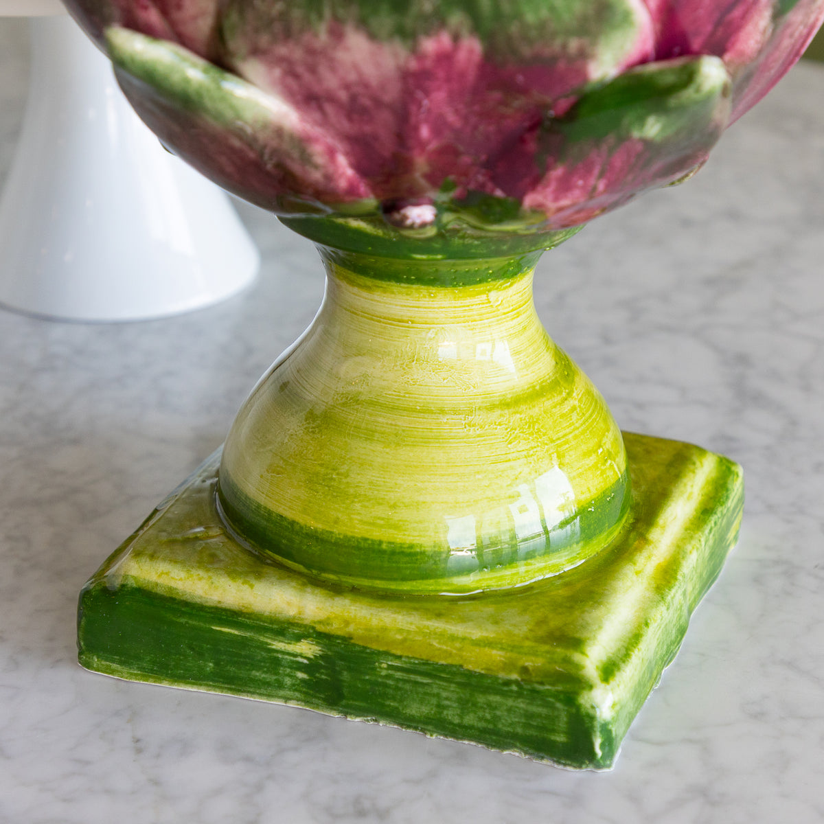 A close-up of a ceramic artichoke-shaped pedestal planter with a glossy green and yellow glazed stem and base, and pink and green glazed petals at the top, set on a light marble surface.