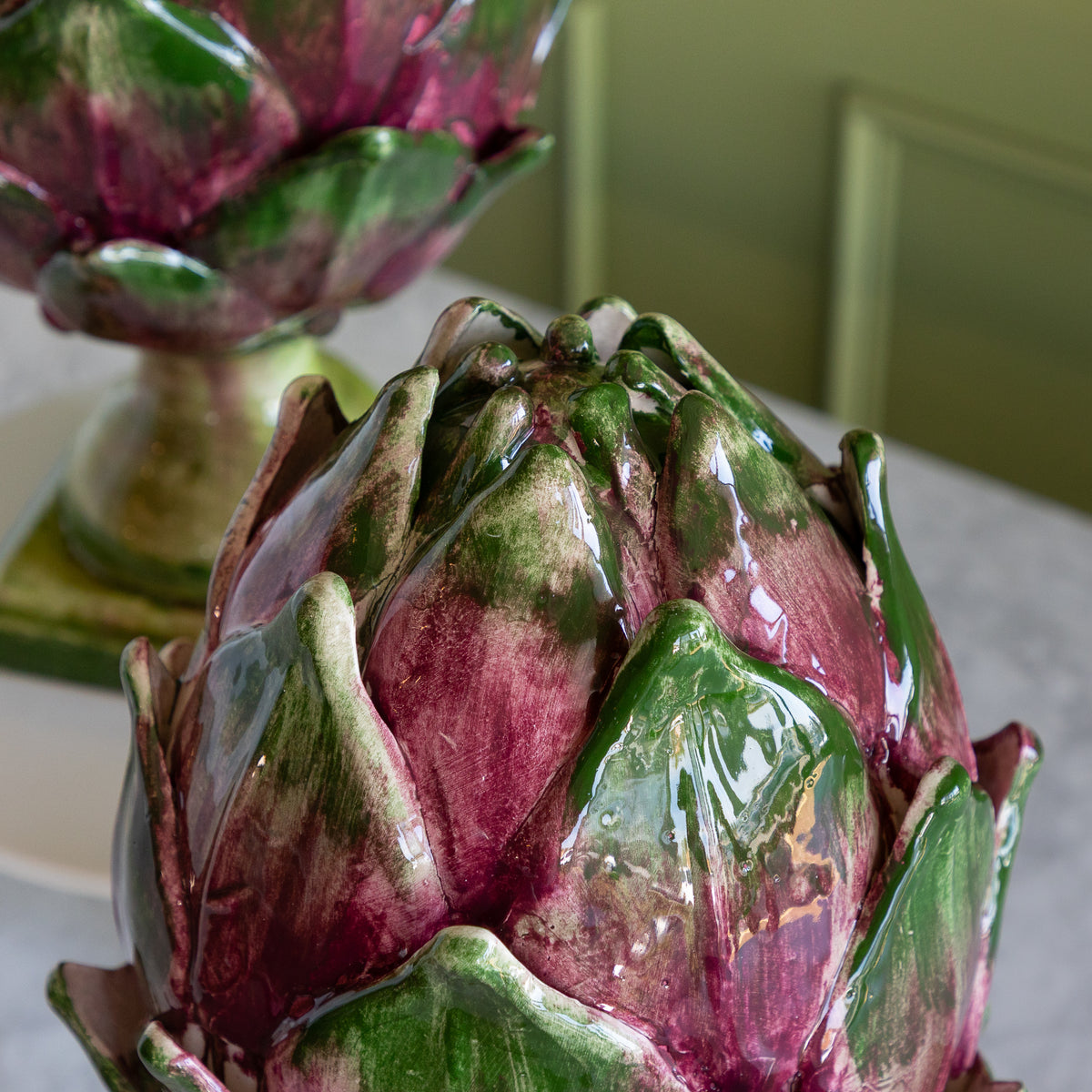 A glossy ceramic artichoke decoration with deep green and burgundy glazed petals, shown close up on a tabletop with another similar piece blurred in the background.