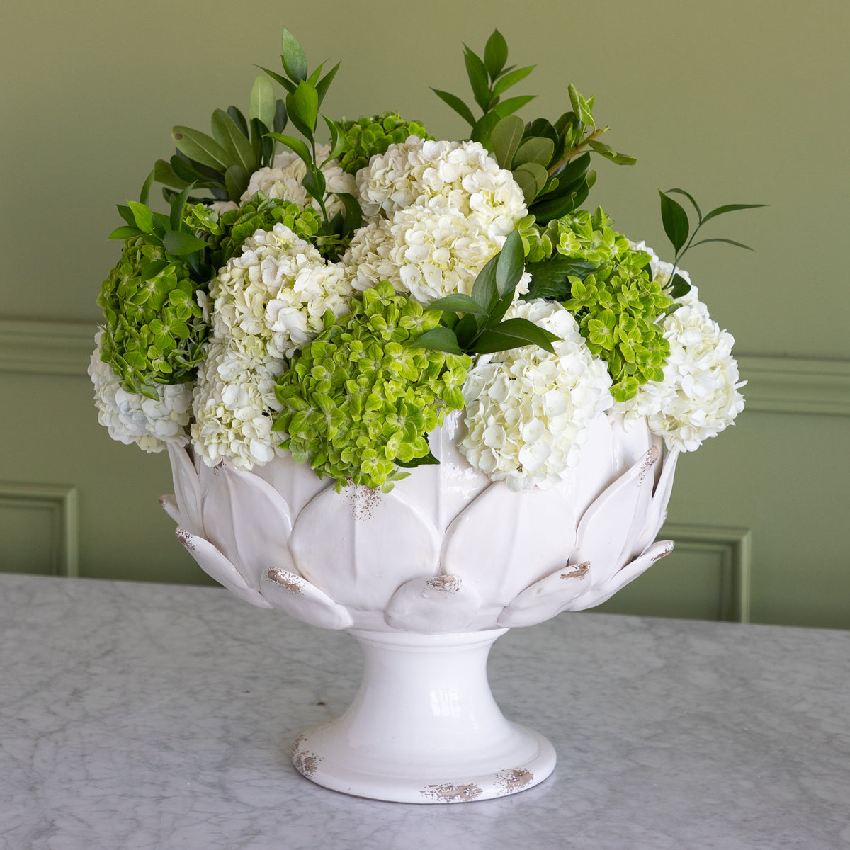 A white ceramic pedestal bowl filled with white and green hydrangea blooms and leafy greenery, displayed on a light marble surface against a sage green paneled wall.