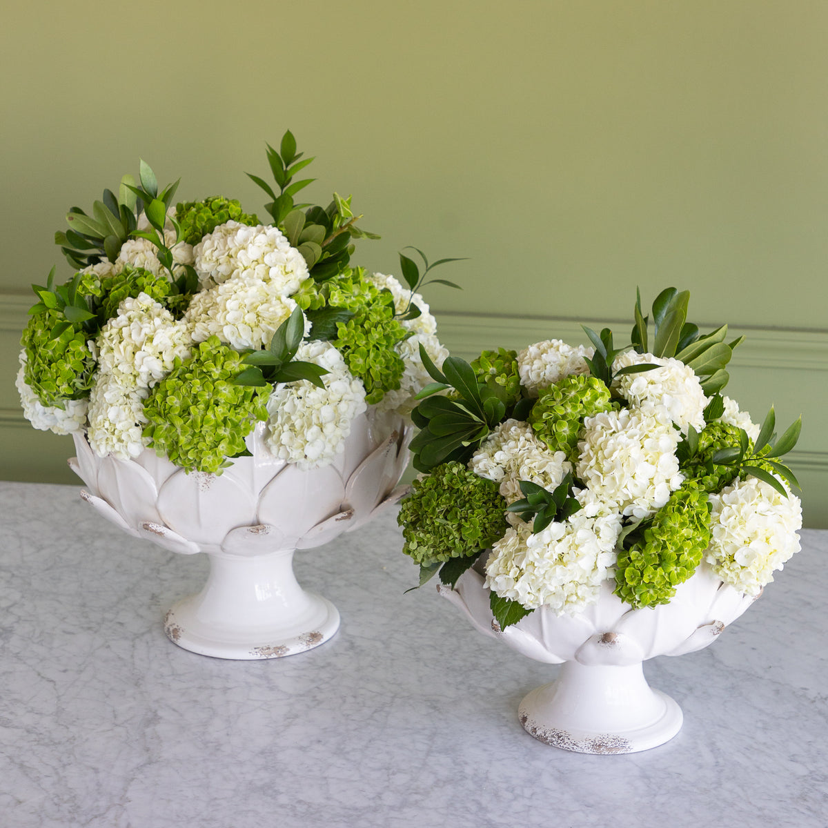 Two white pedestal flower bowls filled with white and green hydrangea bouquets, displayed on a light marble surface against a sage green wall.