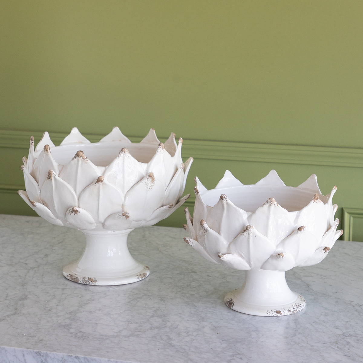 Two white ceramic artichoke-shaped pedestal bowls with distressed gold detailing, displayed on a light gray marble surface against a sage green paneled wall.