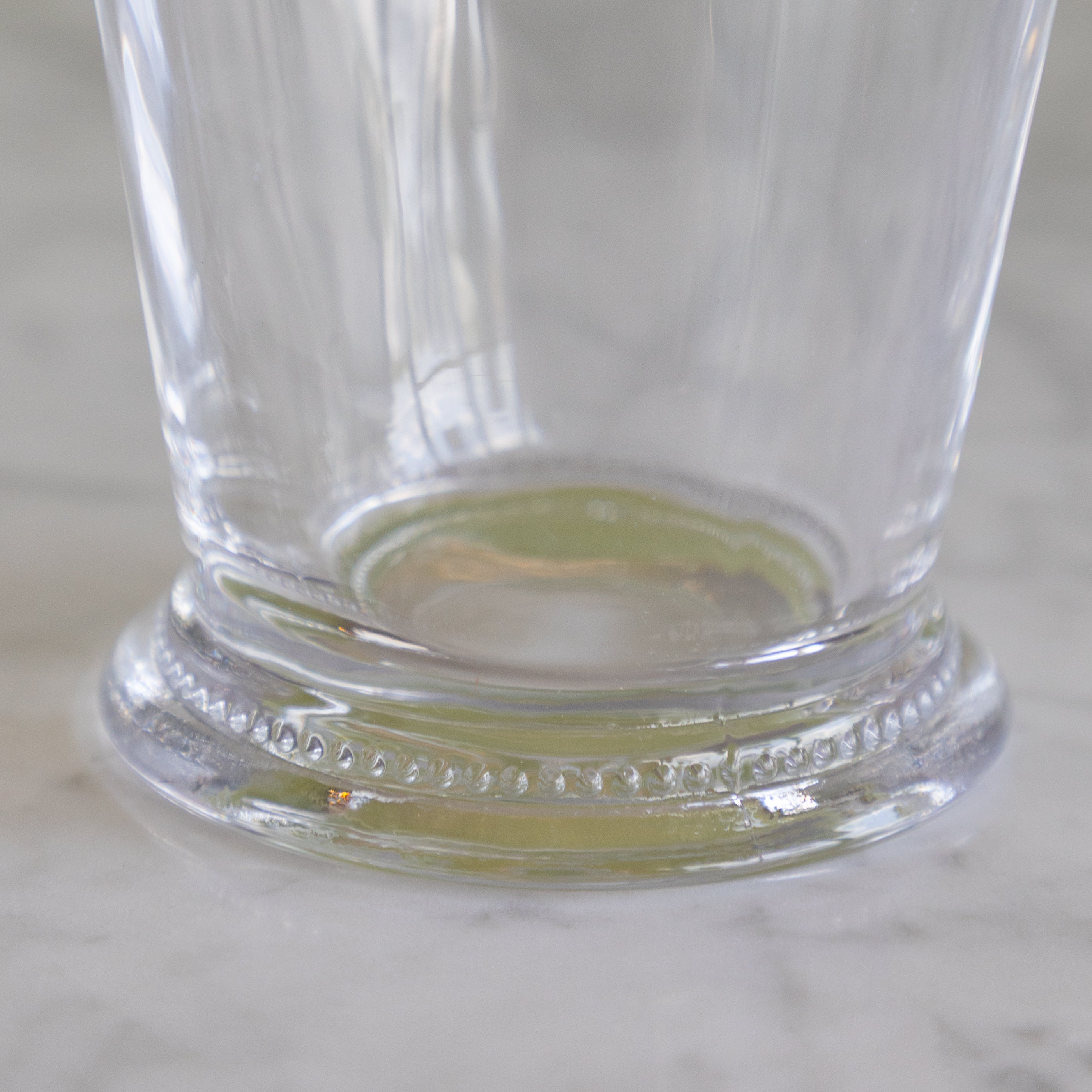 A close-up of a clear glass drinking cup with a decorative beaded base, resting on a light marble surface.