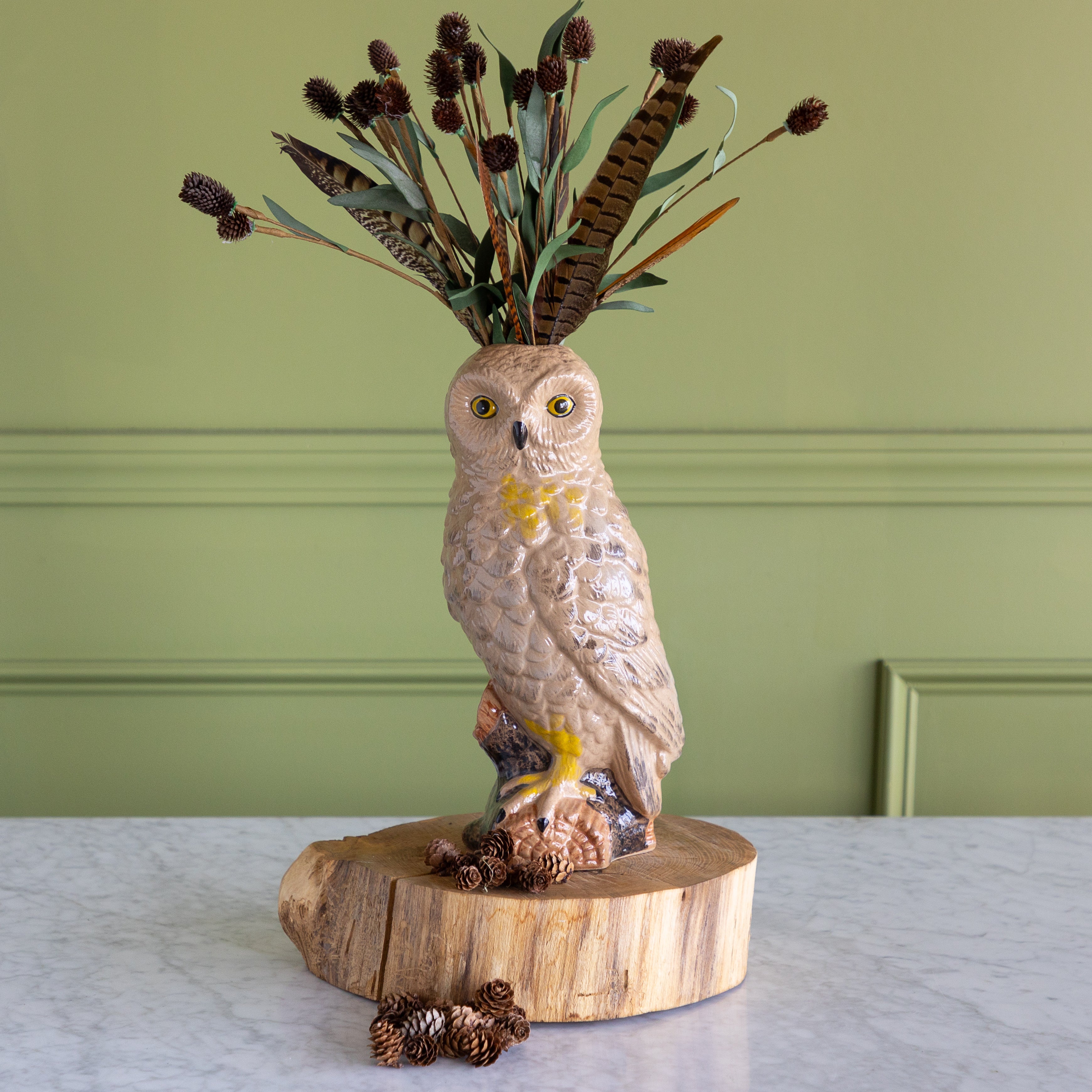 A ceramic owl figurine with yellow eyes and a yellow chest, perched on a round wood slice base and holding dried grasses and feathers above its head. Small pinecones rest on the base, set against a sage green paneled wall and light marble surface.