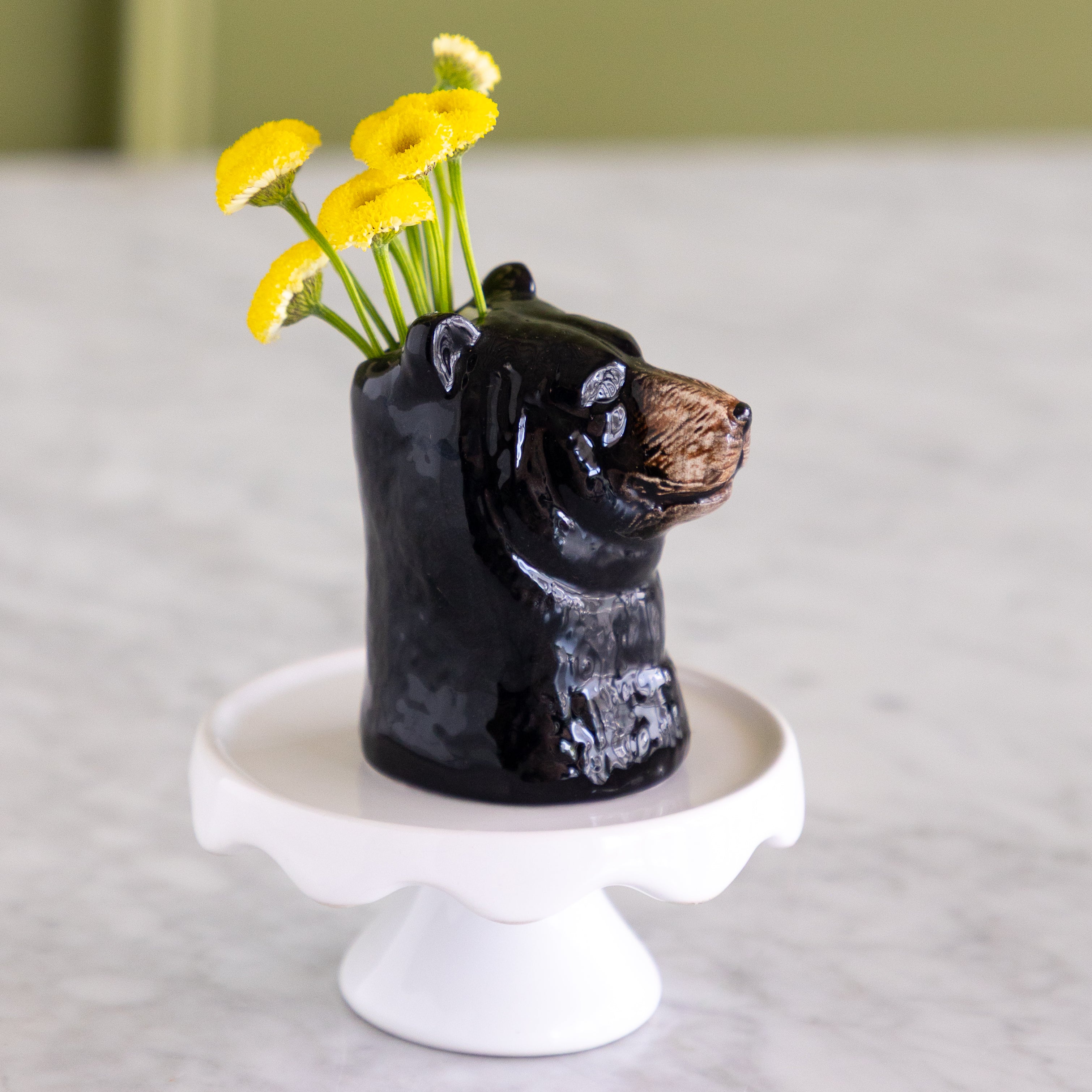 A glossy black bear-shaped ceramic vase holding yellow daisy-like flowers, displayed on a white pedestal stand against a softly blurred kitchen background.