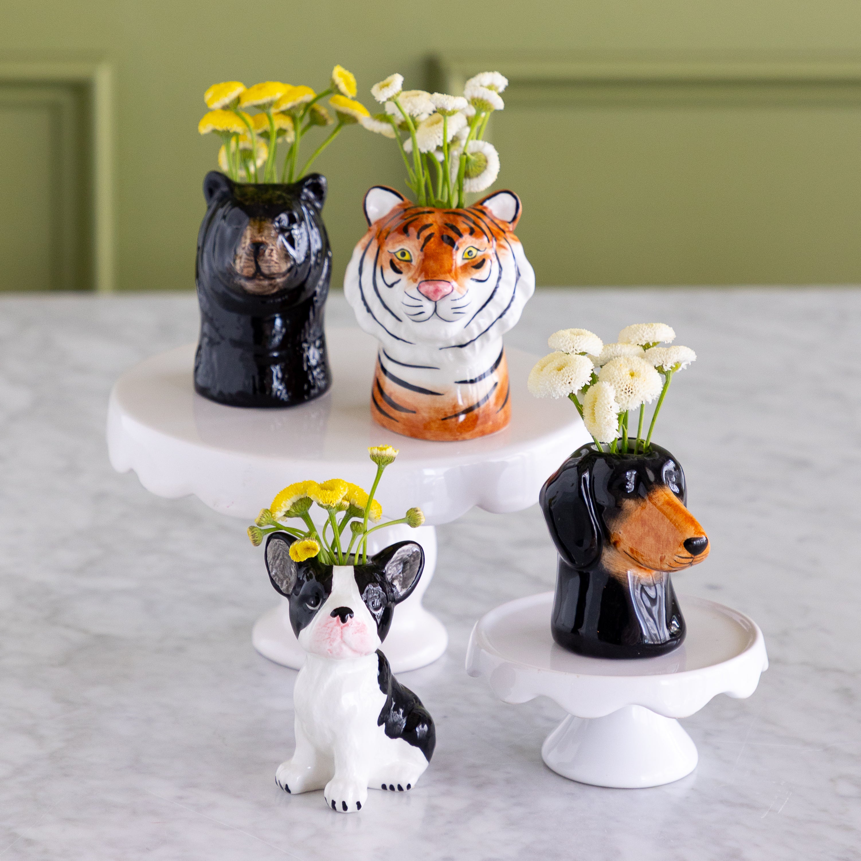 Three ceramic animal-shaped flower vases displayed on white cake stands and a marble surface: a black bear holding yellow flowers, an orange-and-white tiger holding white daisies, and a black-and-white dachshund holding white flowers.