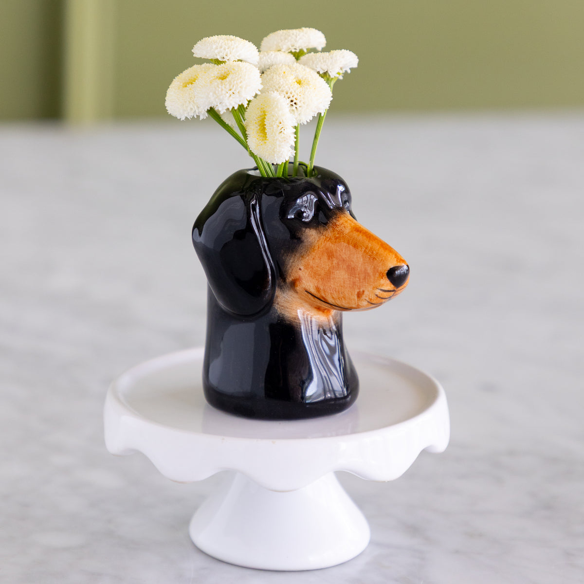 A glossy black ceramic dachshund-shaped vase with a tan face, holding white daisy-like flowers, displayed on a white pedestal stand against a soft-focus indoor background.