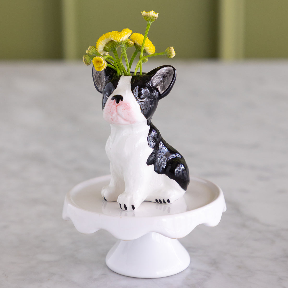 A ceramic French bulldog flower vase sitting on a white pedestal stand, with small yellow flowers placed in its raised head. The vase is photographed on a light marble-like surface with a softly blurred olive-green background.