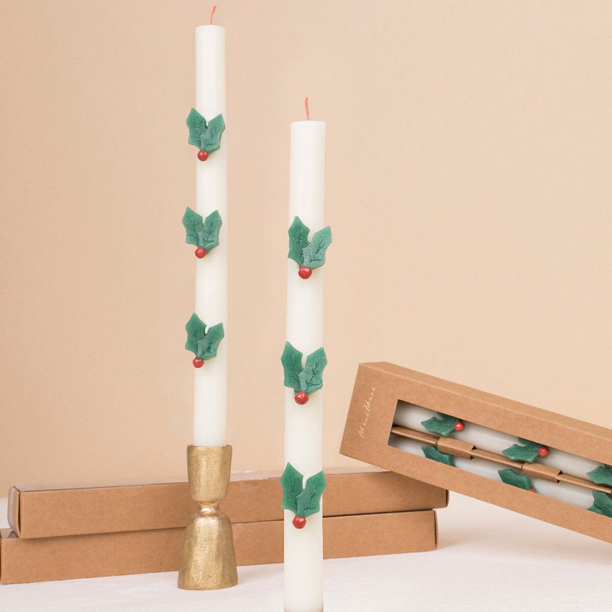 Two white taper candles decorated with green holly leaves and small red berries, shown with a gold candlestick holder and kraft gift boxes on a beige background.