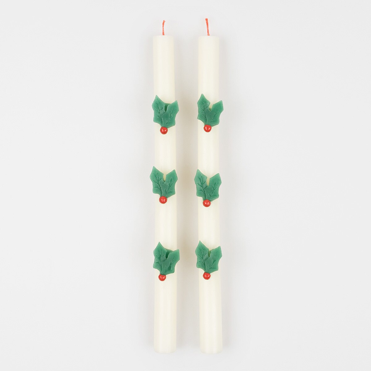 Two white taper candles decorated with green holly leaves and small red berries, shown upright on a plain light gray background.