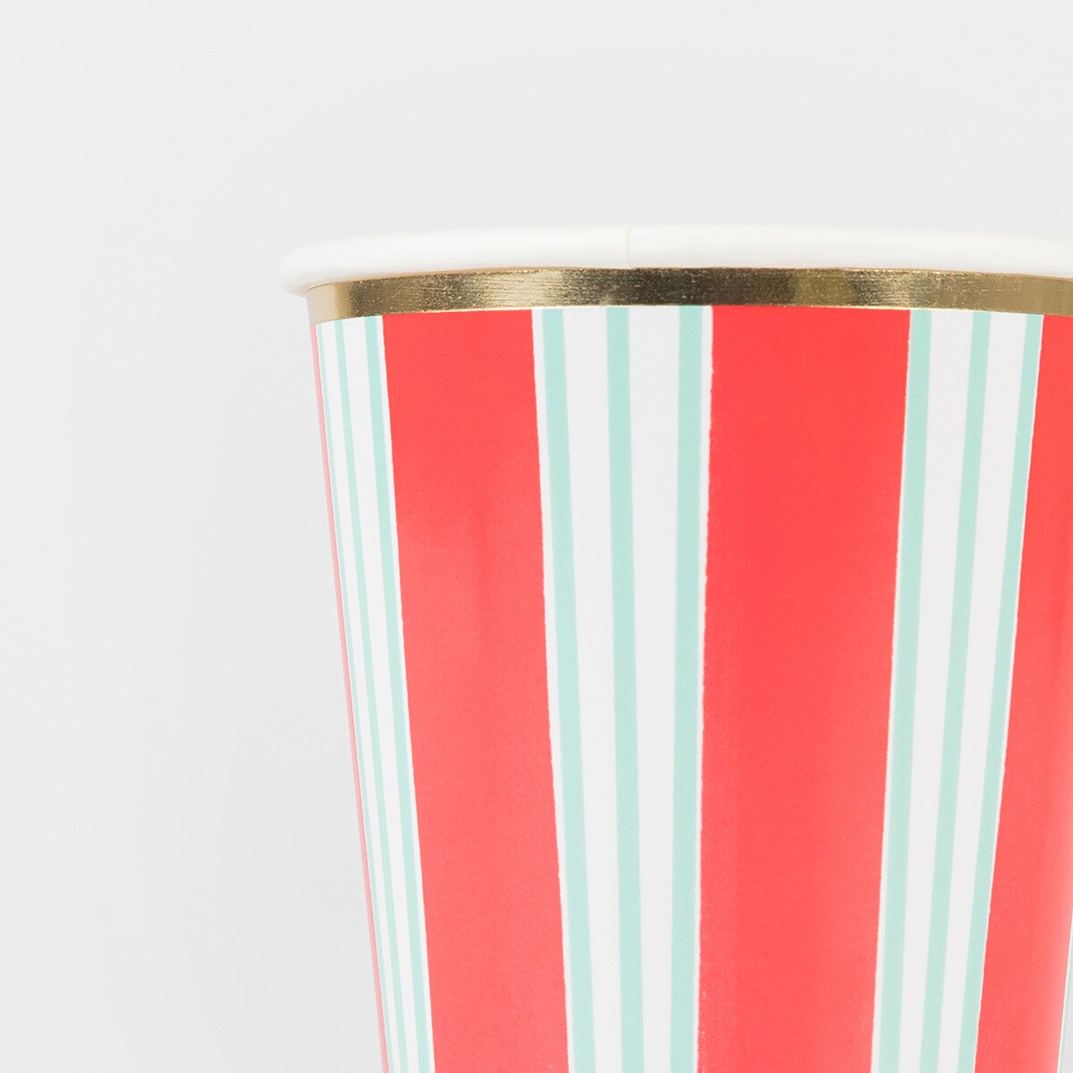 A close-up of a paper cup with vertical red, white, and light blue stripes, featuring a metallic gold rim and a white top edge, set against a plain light gray background.
