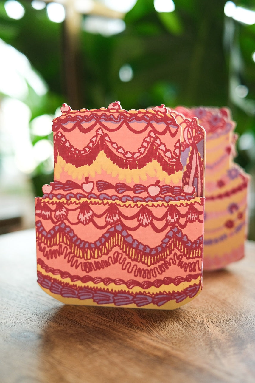 Decorative pink cake-shaped greeting card on a wooden surface with a blurred green background.