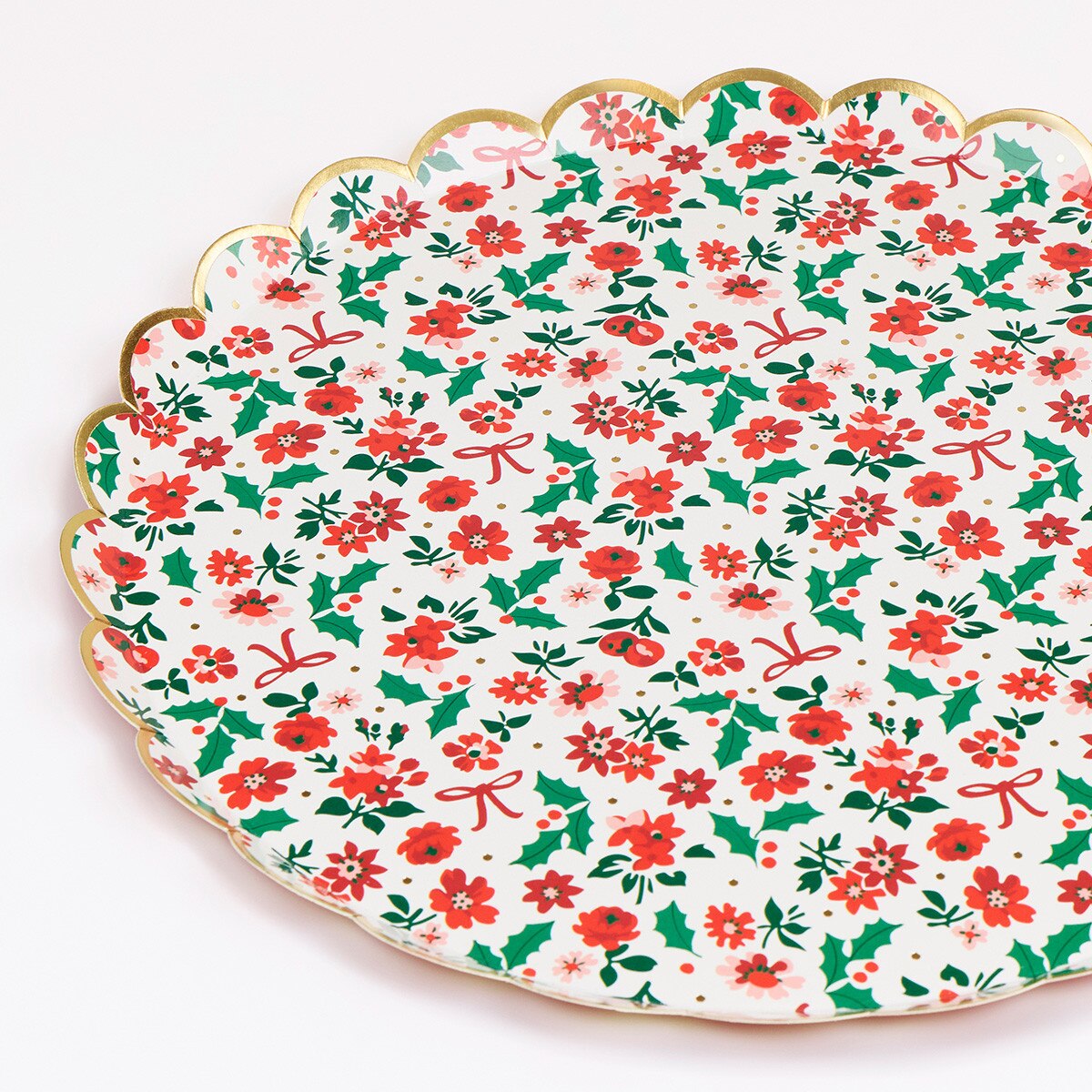A scalloped ceramic plate with a festive Christmas floral pattern featuring red flowers, green holly leaves, and red bows on a white background, finished with a gold rim.