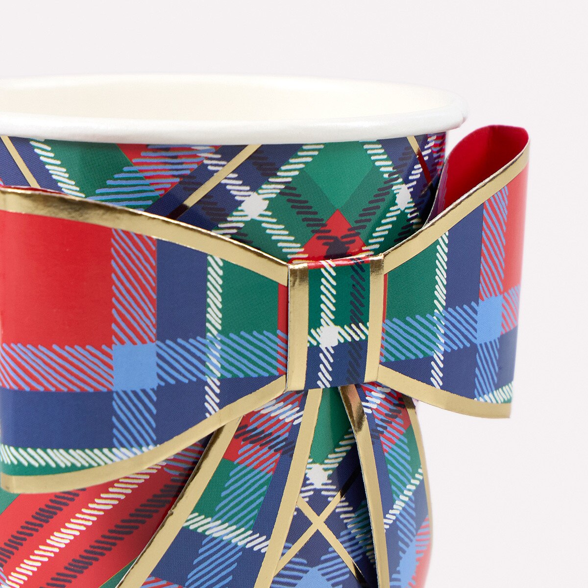 A close-up of a paper cup wrapped in a festive tartan plaid pattern with a large bow. The pattern features red, green, blue, and white plaid sections outlined with gold trim, set against a plain light gray background.
