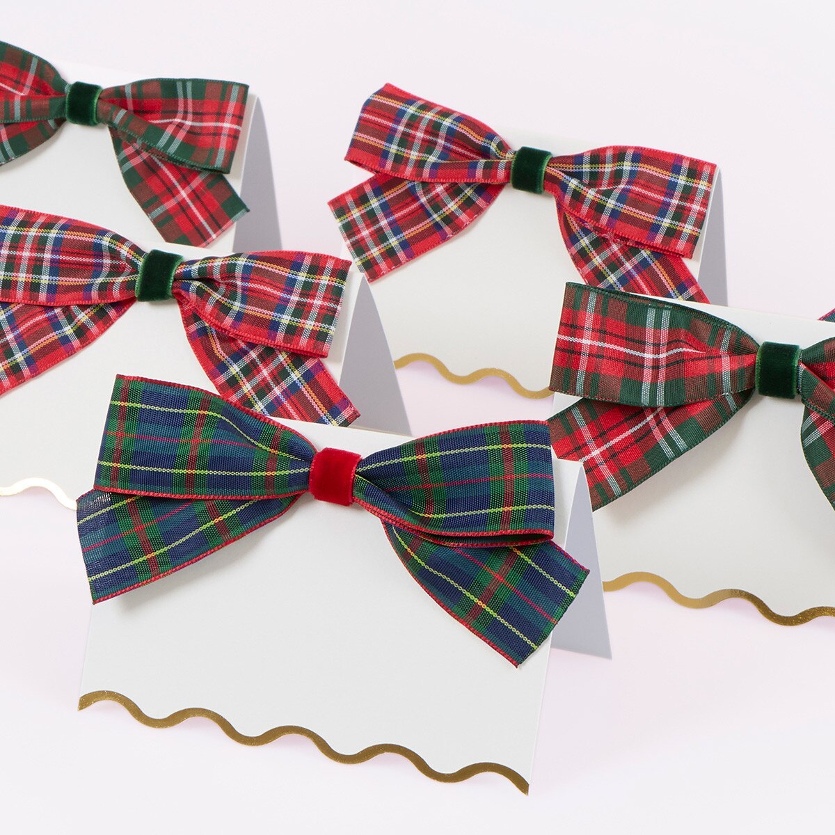 White paper place cards with scalloped edges, each decorated with a plaid ribbon bow. The bows feature red, green, blue, and yellow tartan patterns, accented with small red or green velvet ribbon ties.