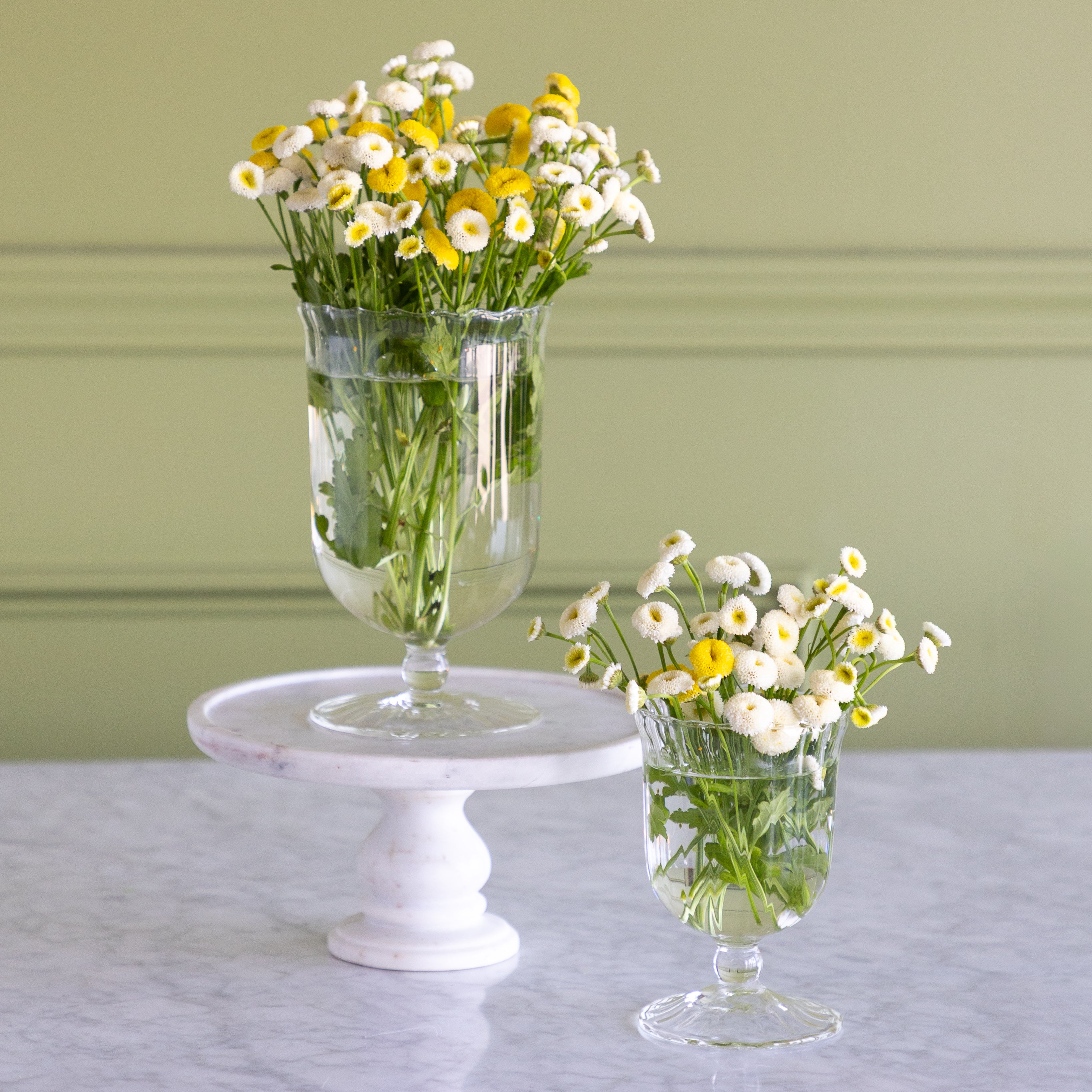 Two clear glass pedestal vases filled with white and yellow daisies, displayed on a white marble cake stand and a light gray marble table against a sage green paneled wall.
