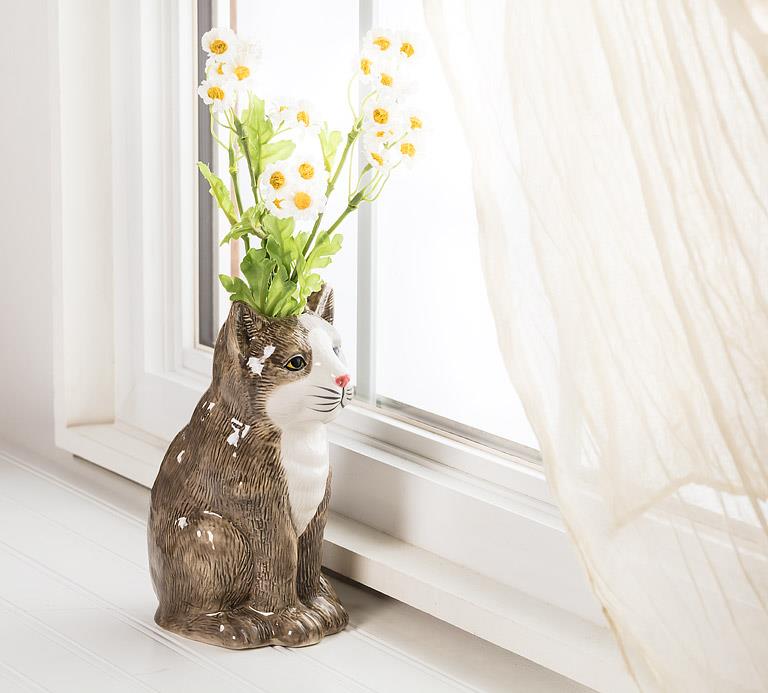 A brown and white cat shaped ceramic vase sitting on a surface next to a window as if it is gazing outside.