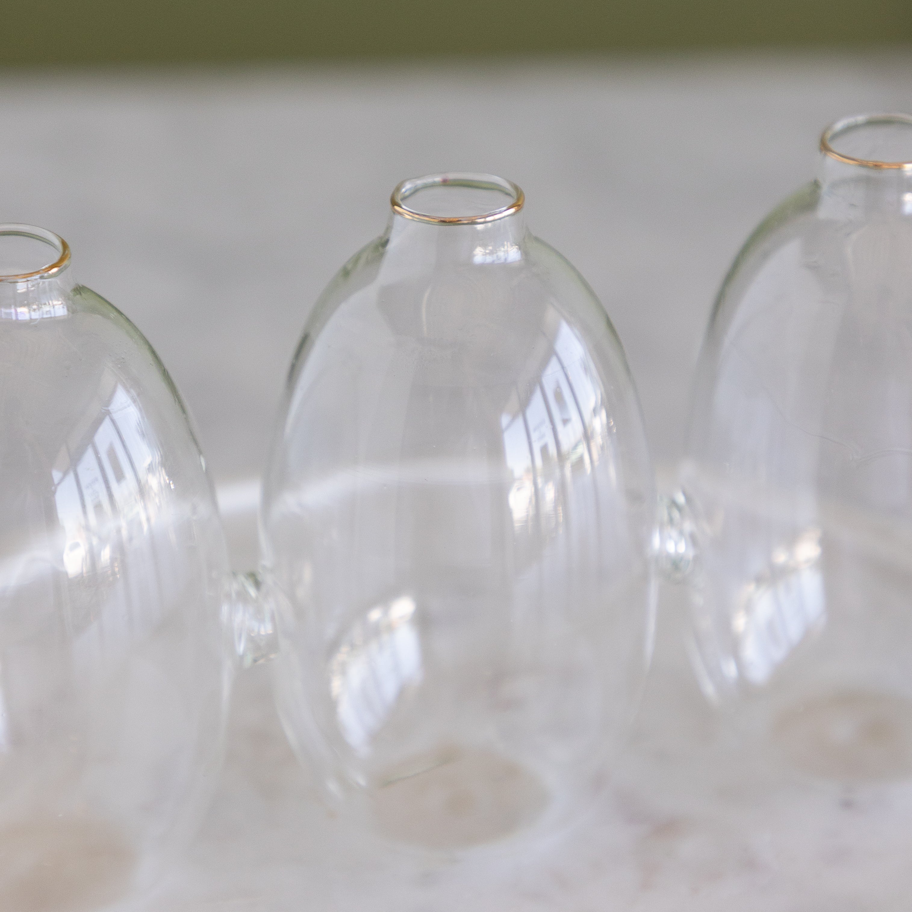 Three clear glass bud vases with small gold rims at the top, arranged on a light marble surface.