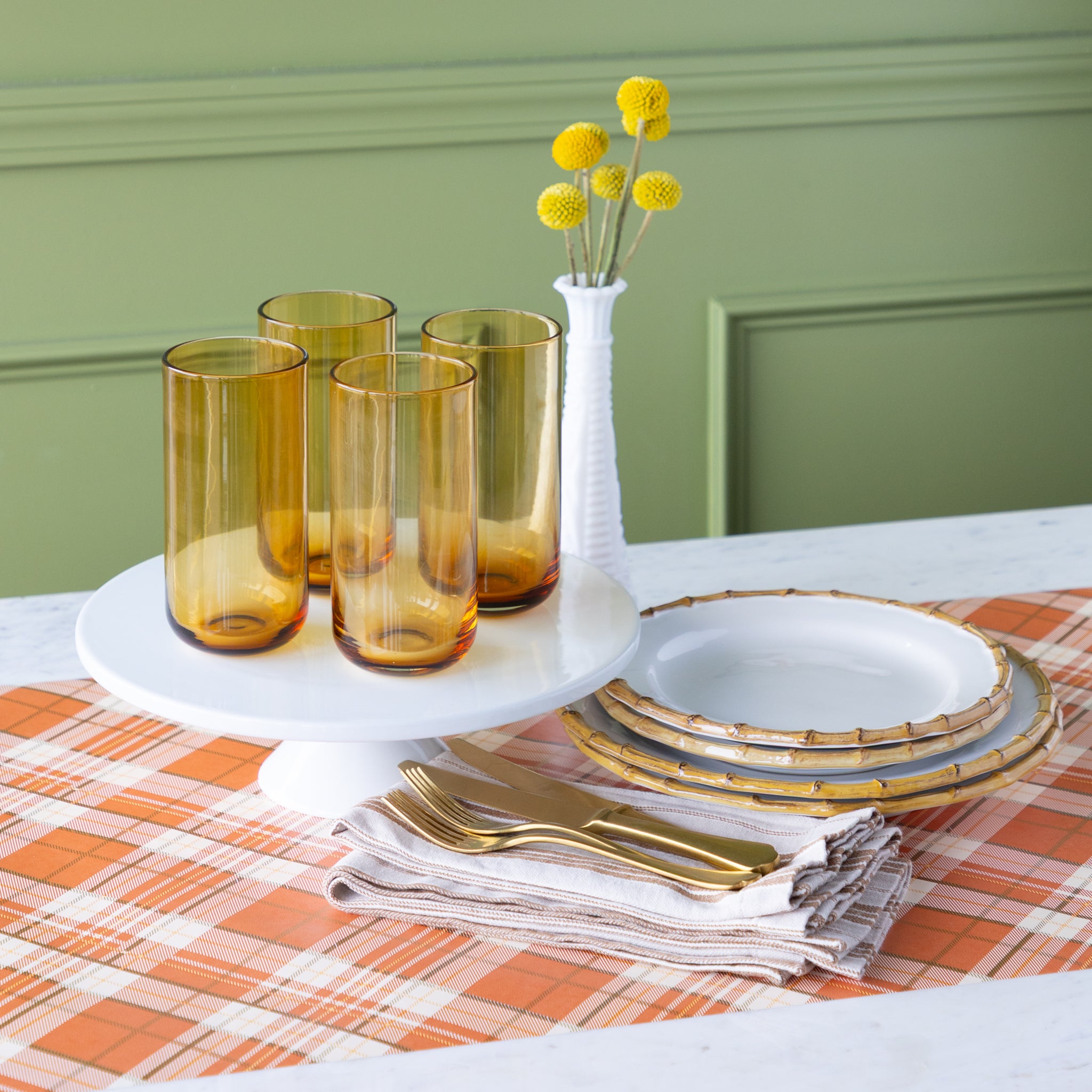 Table setting with amber glasses, gold cutlery, Juliska Bamboo Dinner Plates and a vase with yellow flowers on the Orange Plaid Runner.