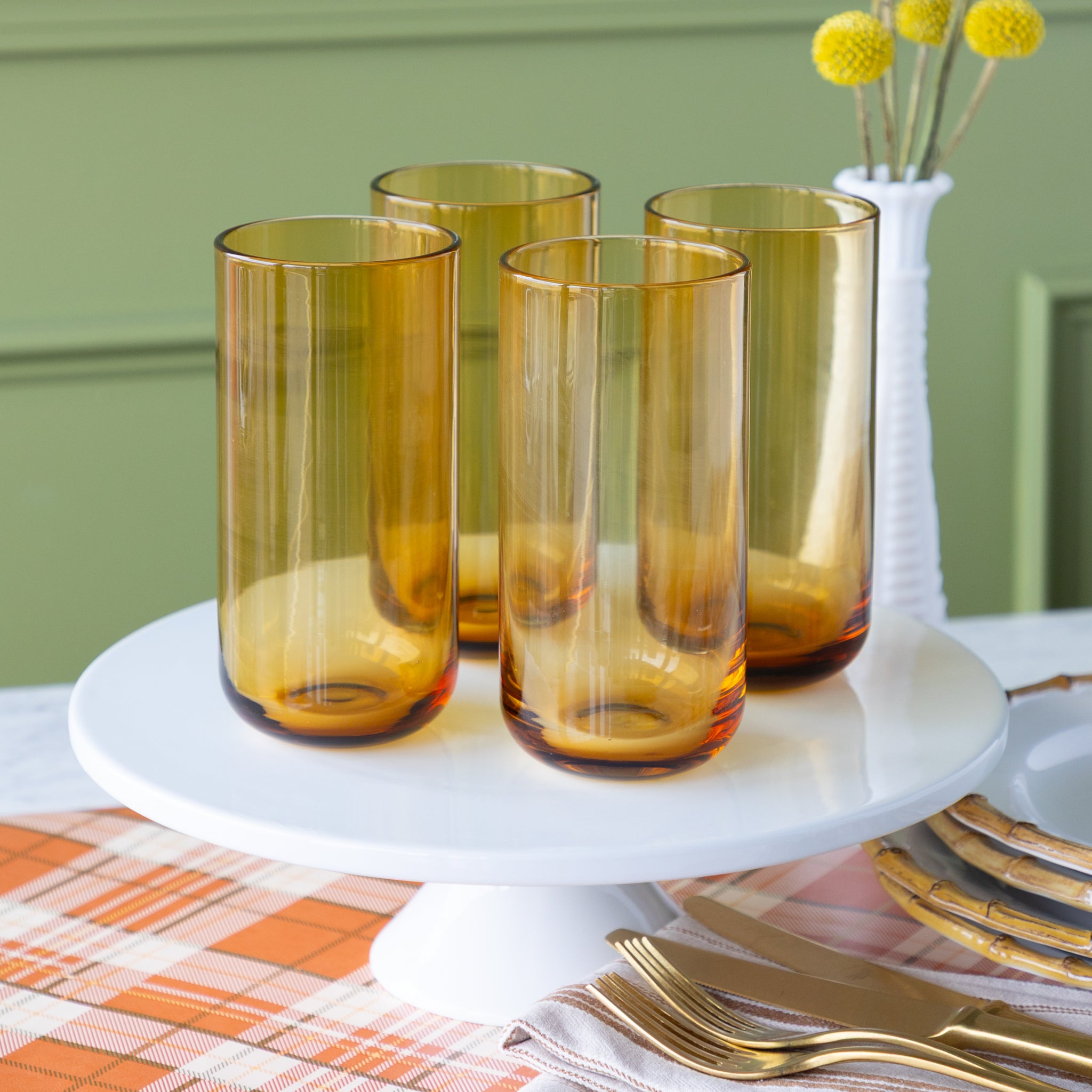 Set of four amber-colored glasses on a white cake stand with a green wall and table setting in the background.