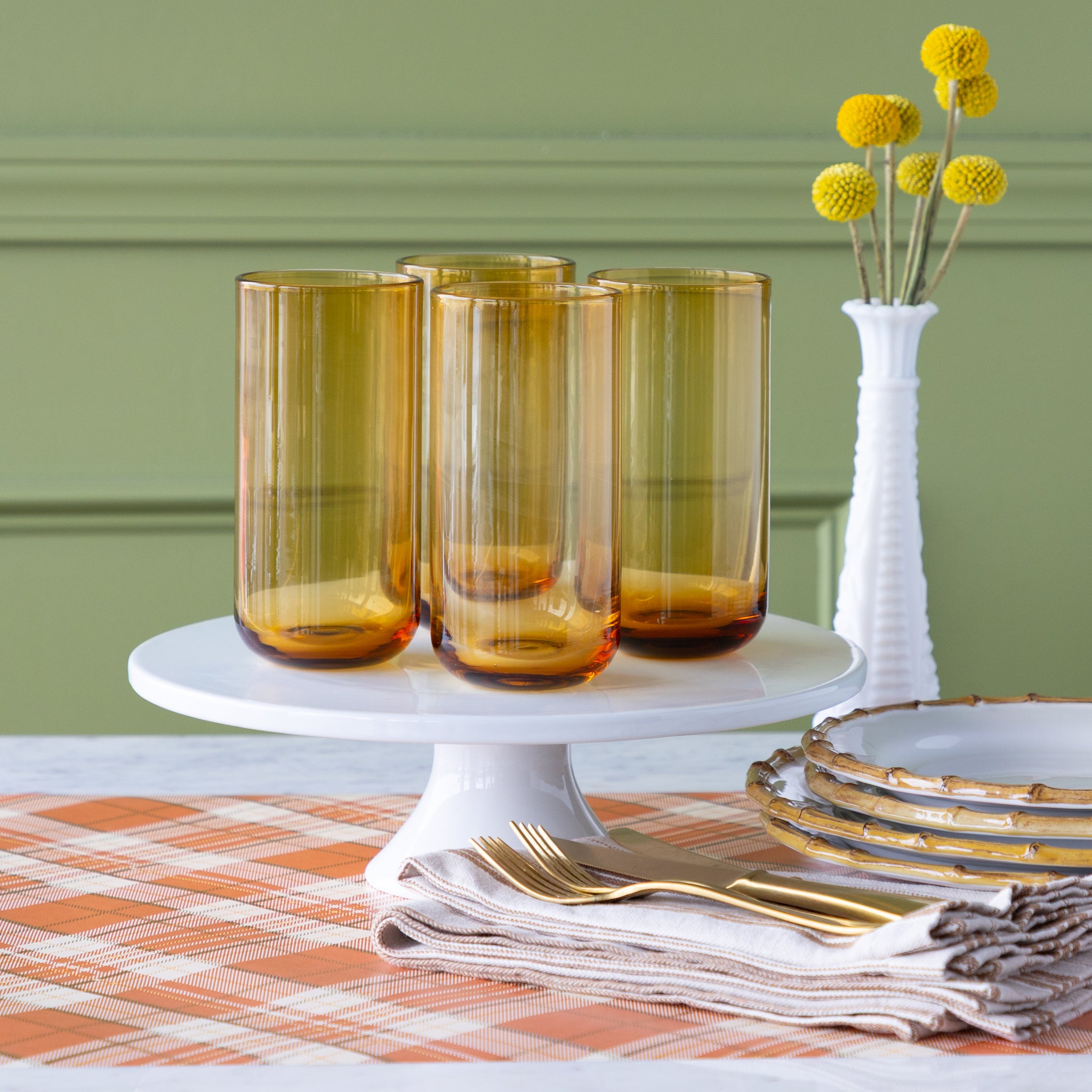 Set of four amber-colored glasses on a white cake stand on the Orange Plaid Runner and decorative elements.