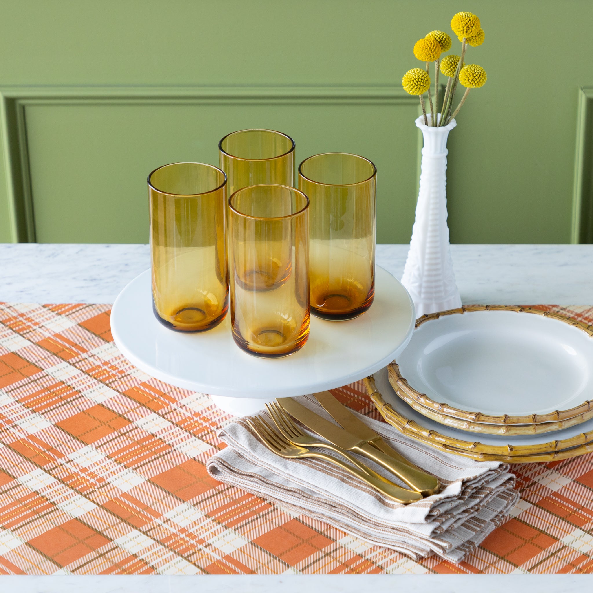 Table setting with amber glasses, gold cutlery, Juliska Bamboo Dinner Plates and a vase with yellow flowers on the Orange Plaid Runner.