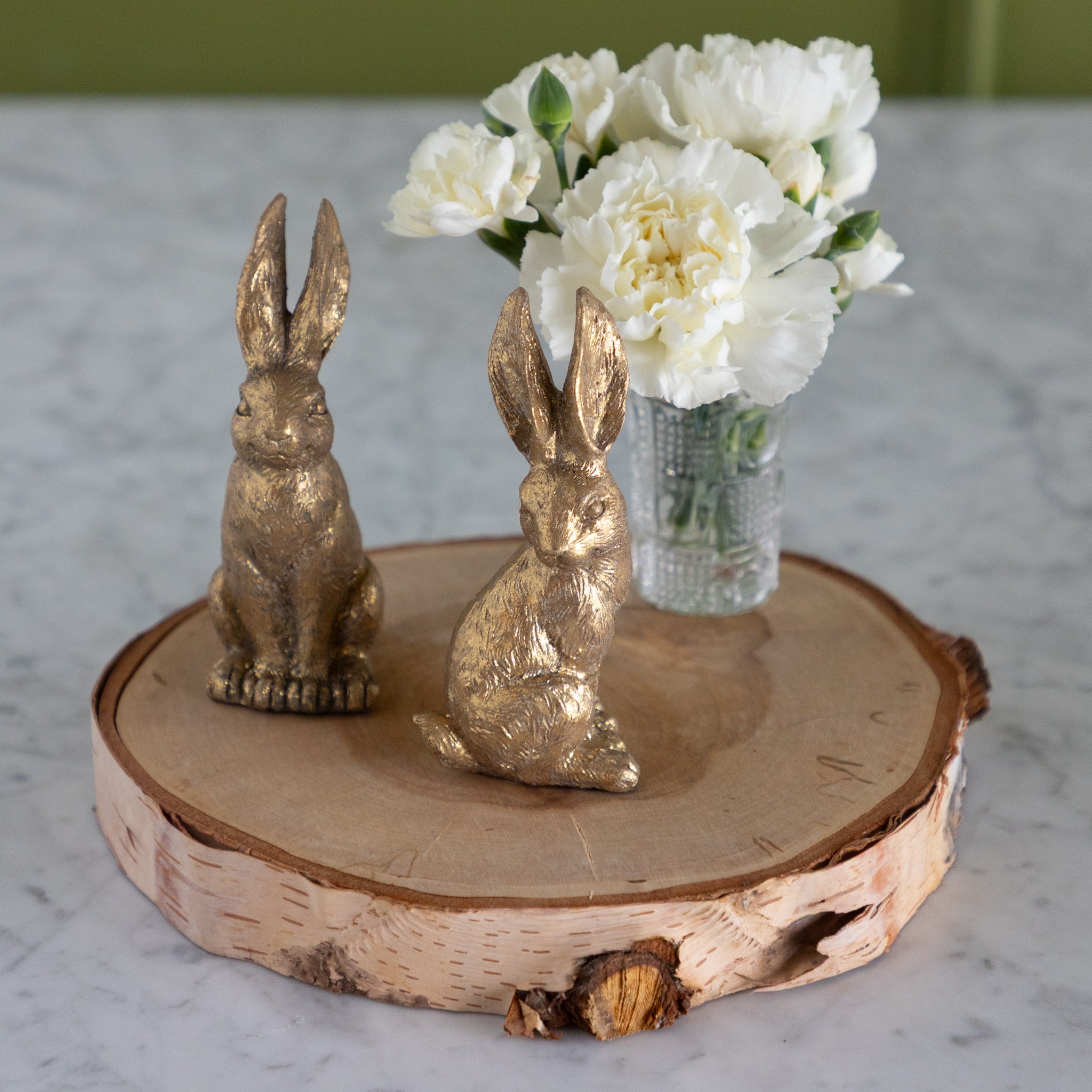 Two gold rabbit figurines on a wooden stand with a vase of white flowers in the background.