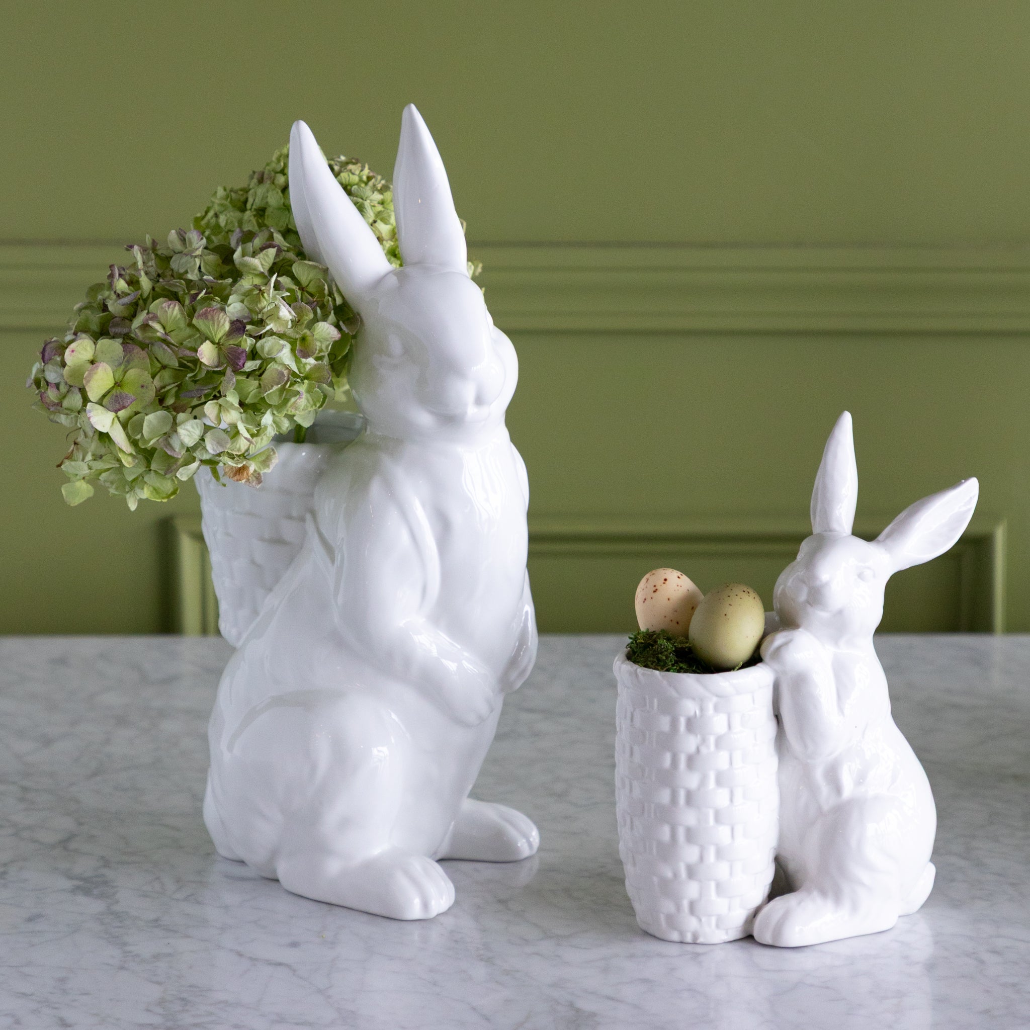 White rabbit-shaped planters with one holding green hydrangeas and the other with Easter eggs on a marble surface.