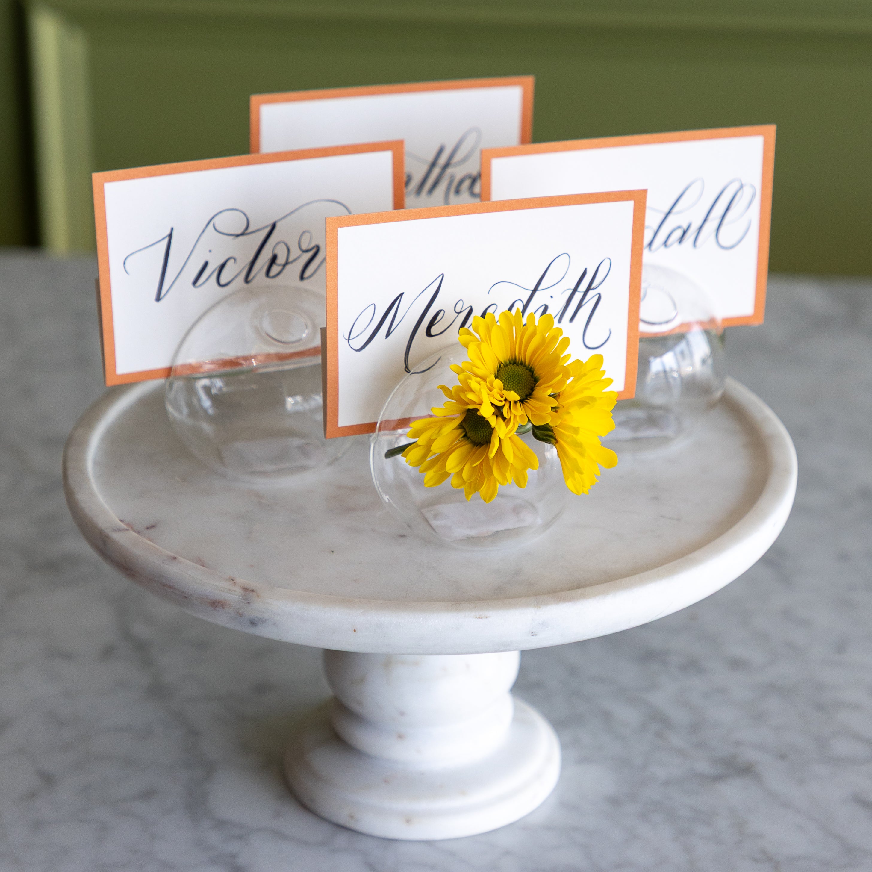 White marble cake stand with yellow flowers and name cards on a marble surface.