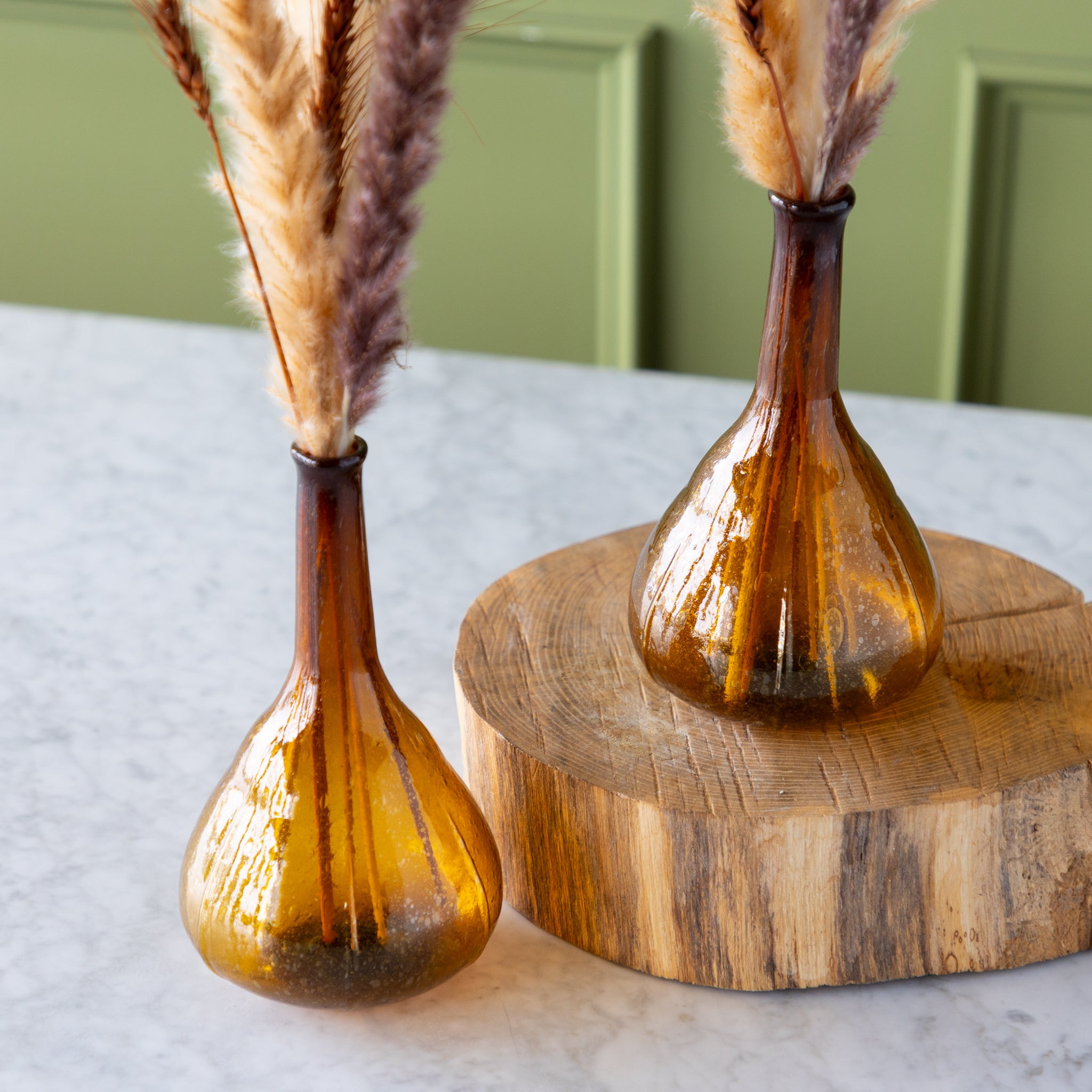 Two amber glass vases with dried pampas grass on a wooden stand against a green wall.