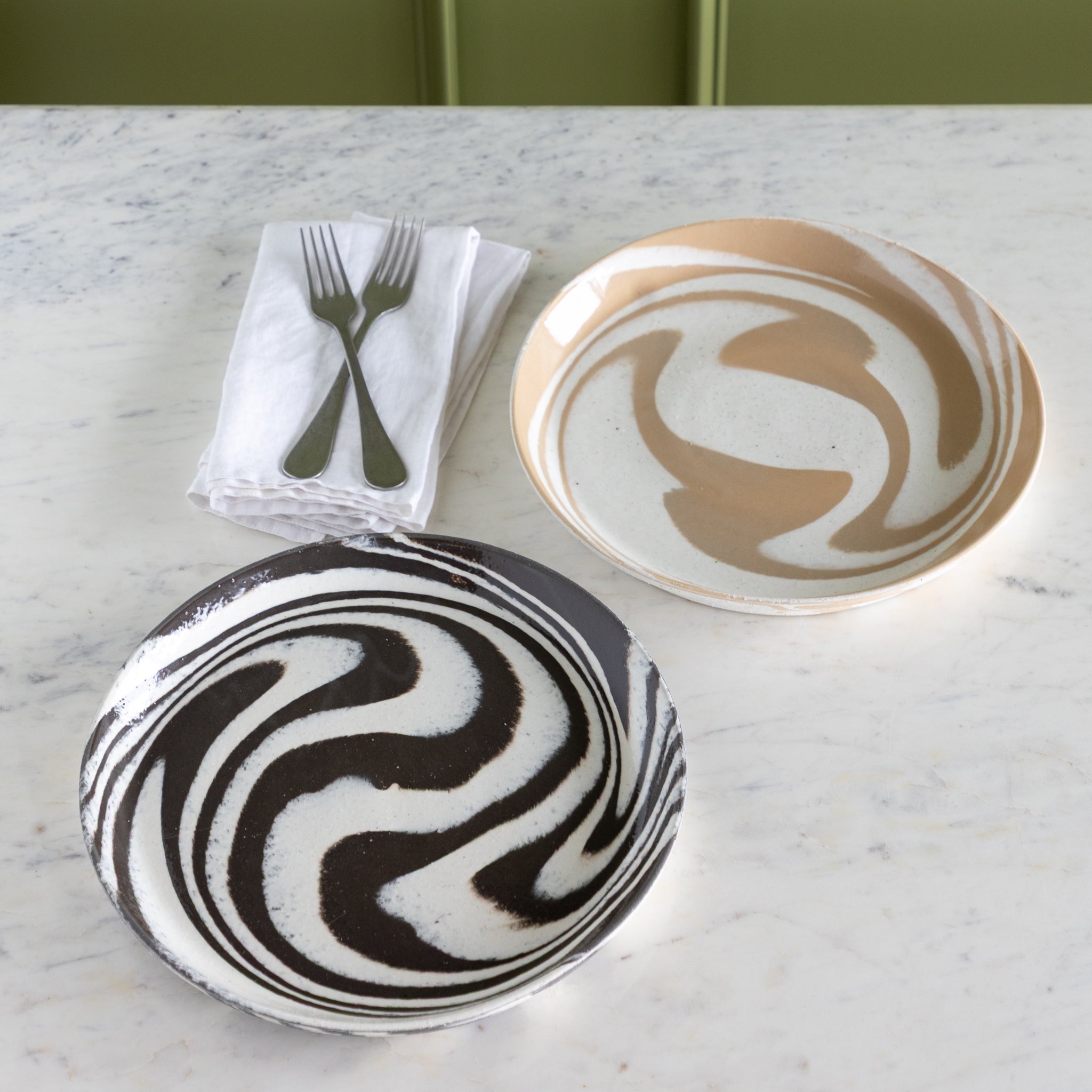 Two ceramic plates with black and white swirl patterns on a marble surface with napkins and cutlery.