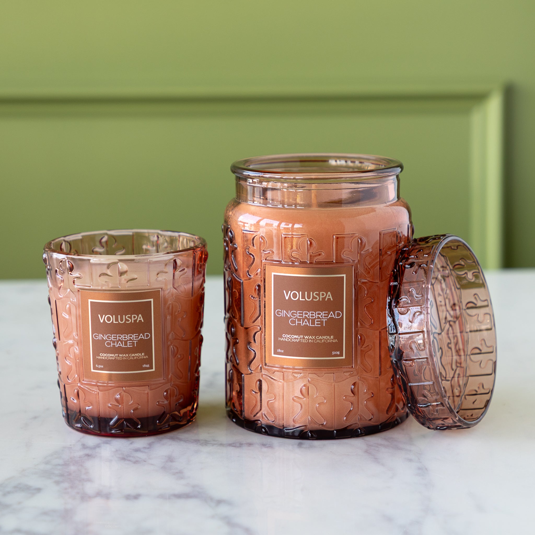 Two Voluspa gingerbread chalet candles on a marble surface with a green background.