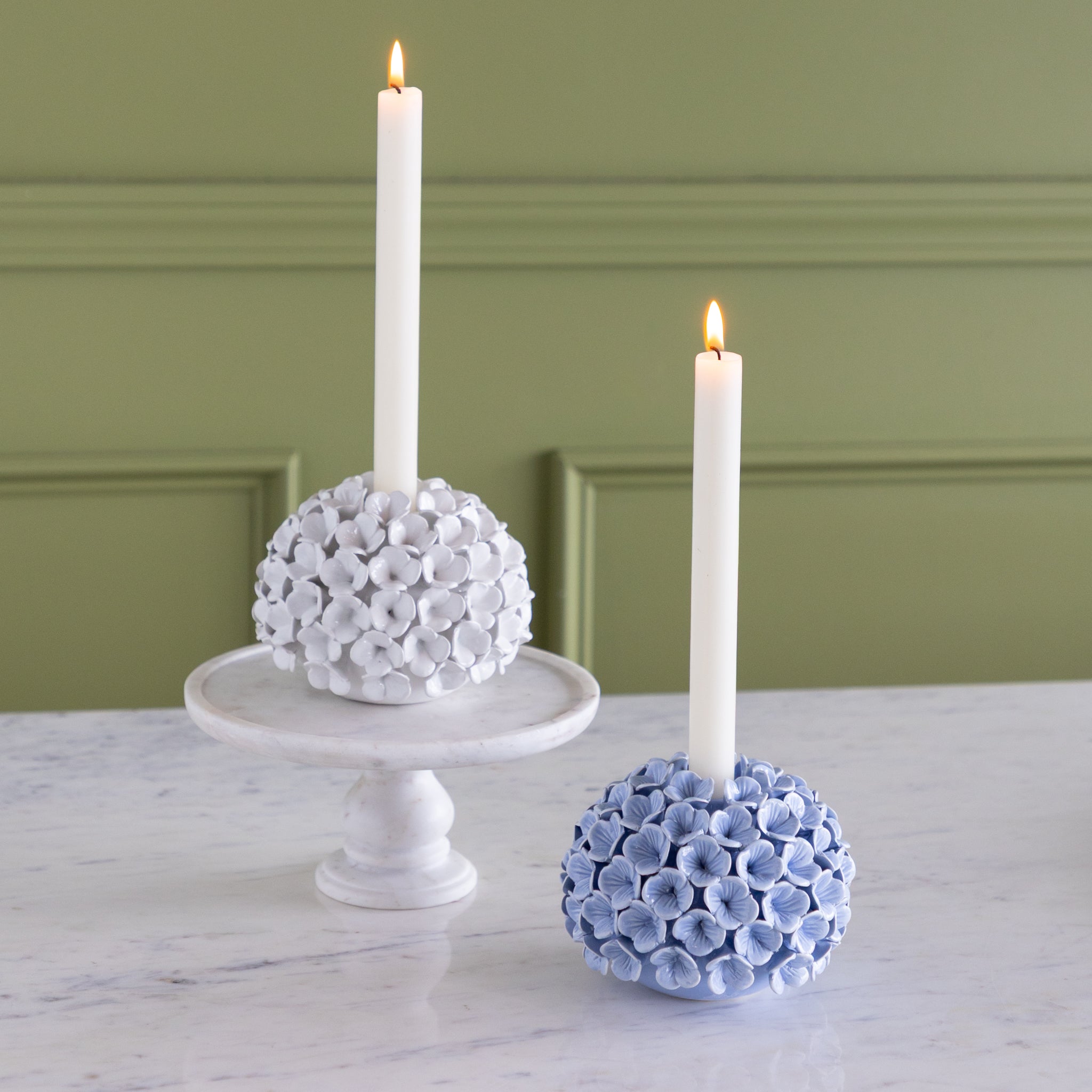 Two candle holders with white candles on a marble surface against a green wall.