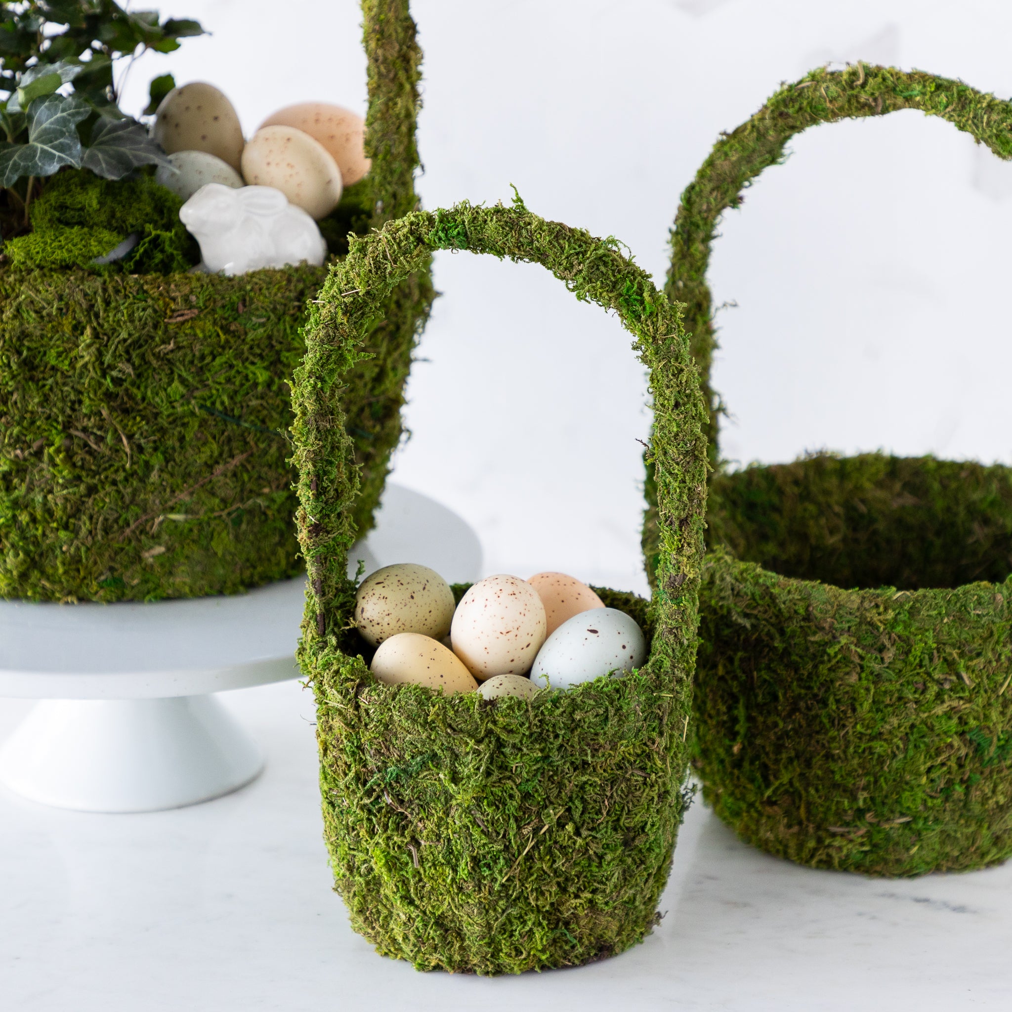 Three decorative moss baskets, one with speckled eggs, another with a floral arrangement and eggs, and an empty larger basket.