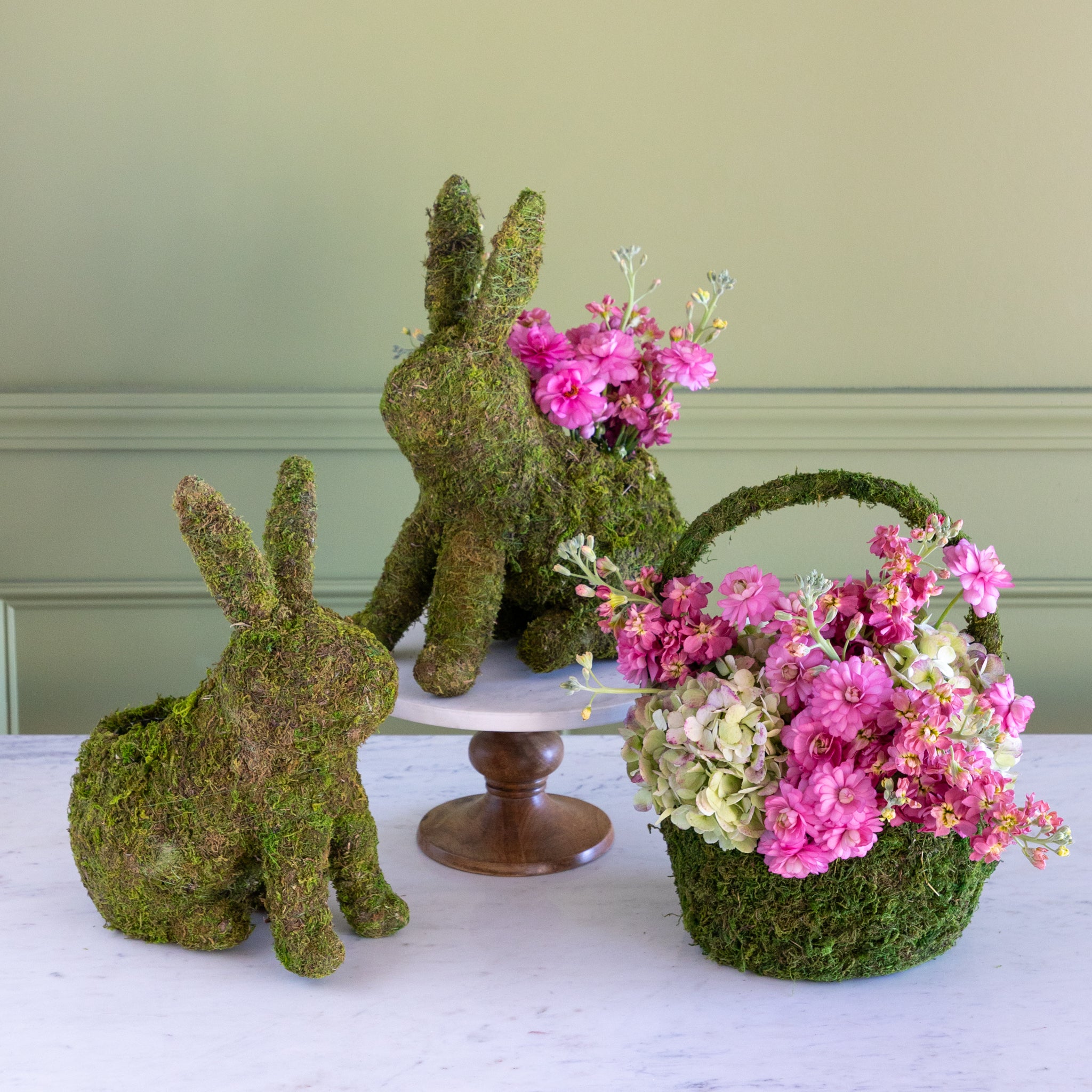 Moss-covered bunny figures and a basket of pink flowers on a white surface with a green wall background.