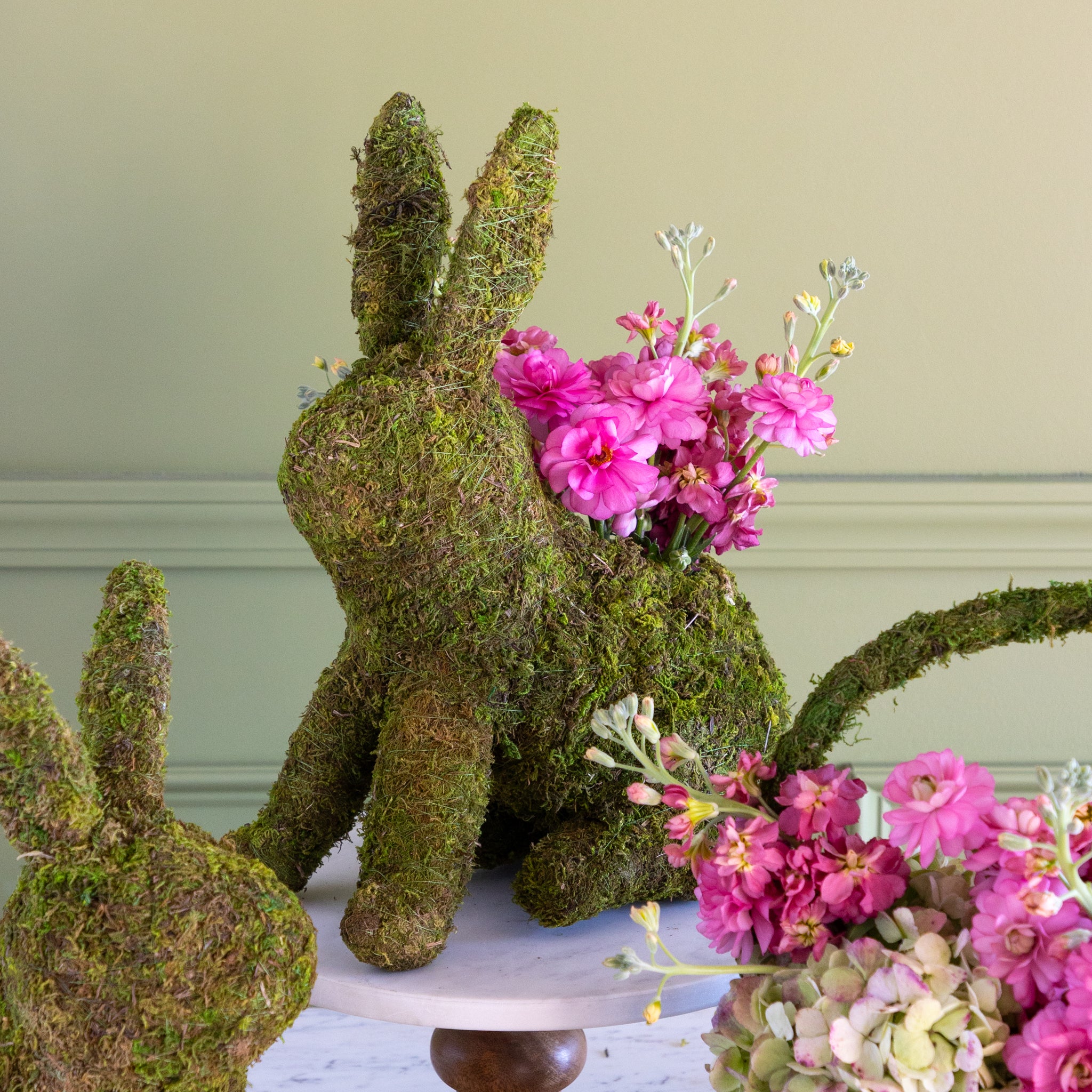 Moss-covered bunny figurine with pink flowers on a decorative stand against a light green wall.